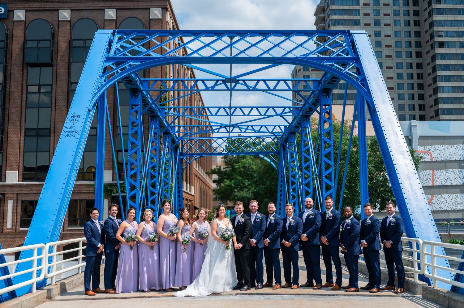 Wedding party poses on a blue bridge in Grand Rapids, bridesmaids in lavender gowns and groomsmen in navy suits.