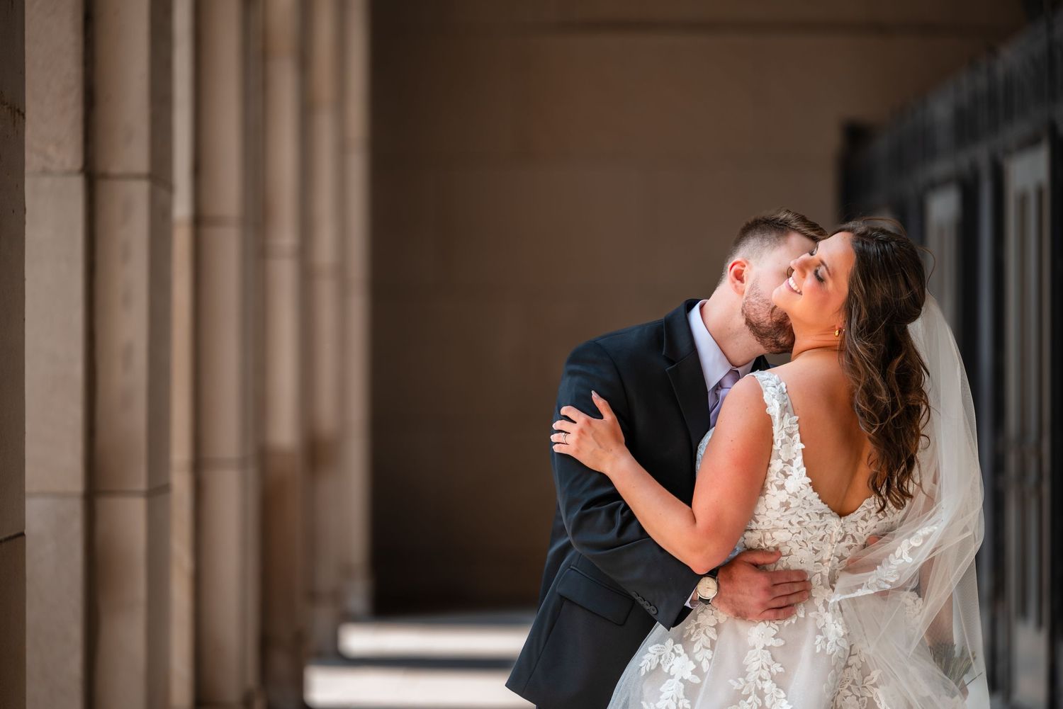 Bride and groom share a romantic embrace between stone columns, with the groom kissing the bride in her lace wedding gown.