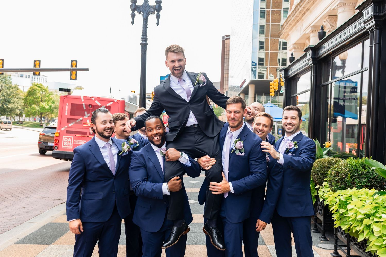 Groomsmen in navy suits lifting groom outdoors on city street.