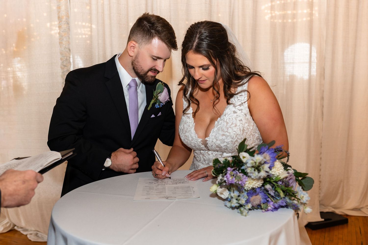 Bride and groom signing wedding documents at a white table, with a blue and white floral bouquet nearby.
