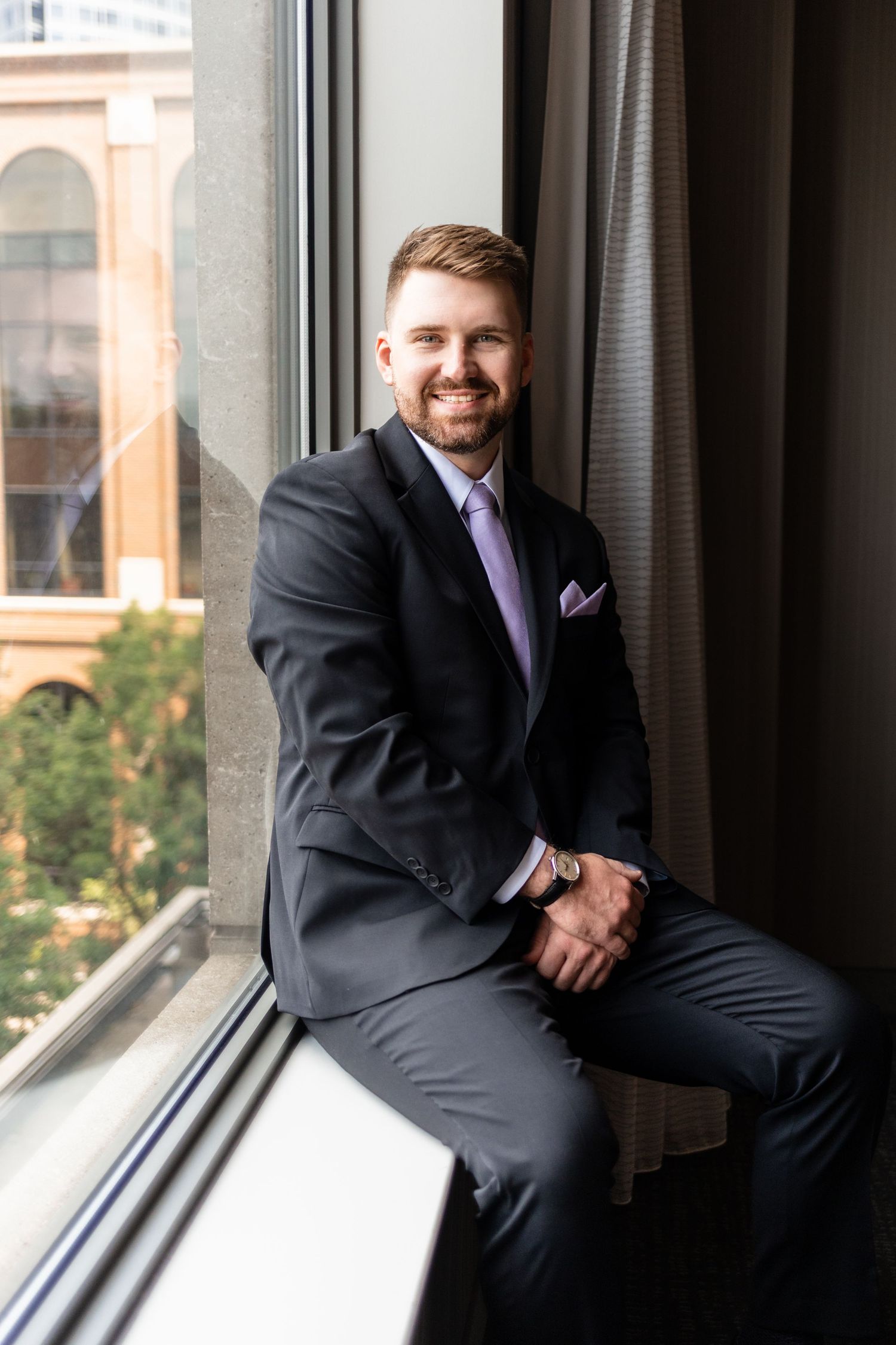 Smiling groom in dark suit with lavender tie sits on windowsill, posing for wedding portrait.