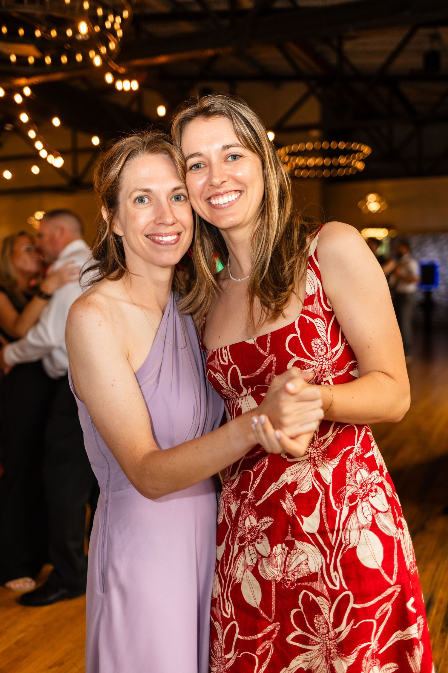 Two smiling women dancing together in elegant dresses at a wedding reception with warm string lights.