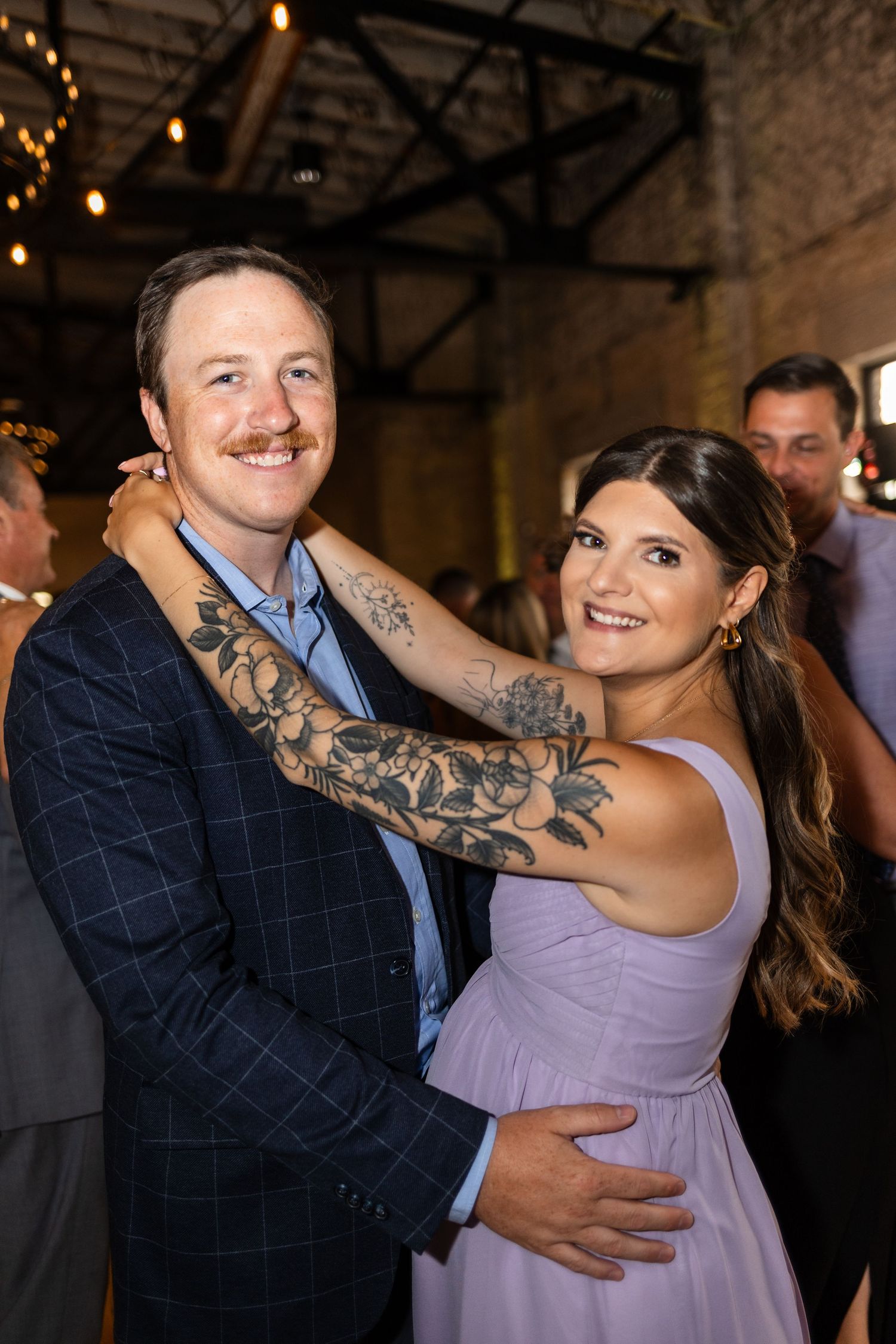 A smiling couple embraces at a wedding reception, man in plaid blazer with floral tattoo sleeve, woman in lavender gown.