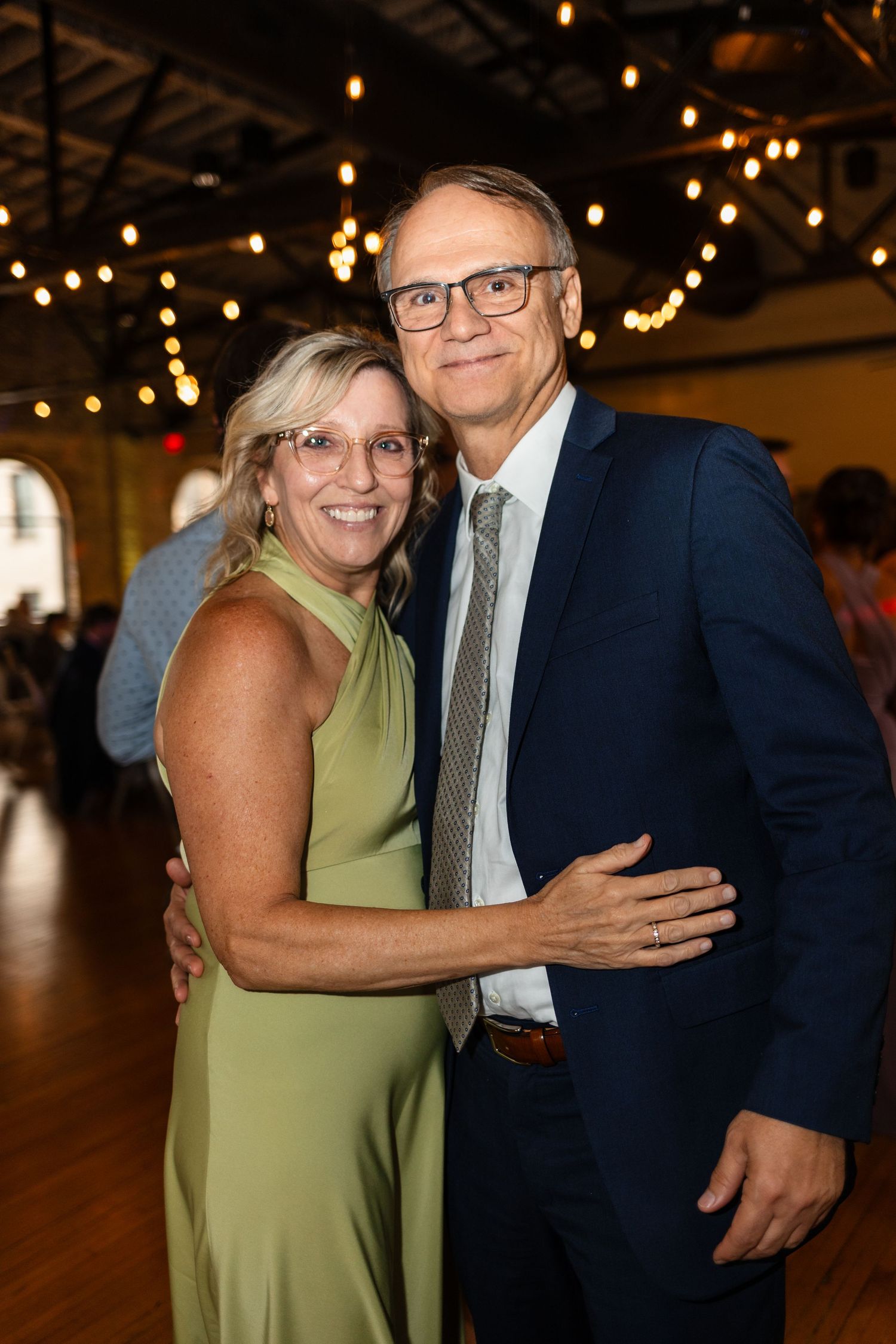 A smiling couple in formal attire poses together at an elegant indoor event with string lights in the background.