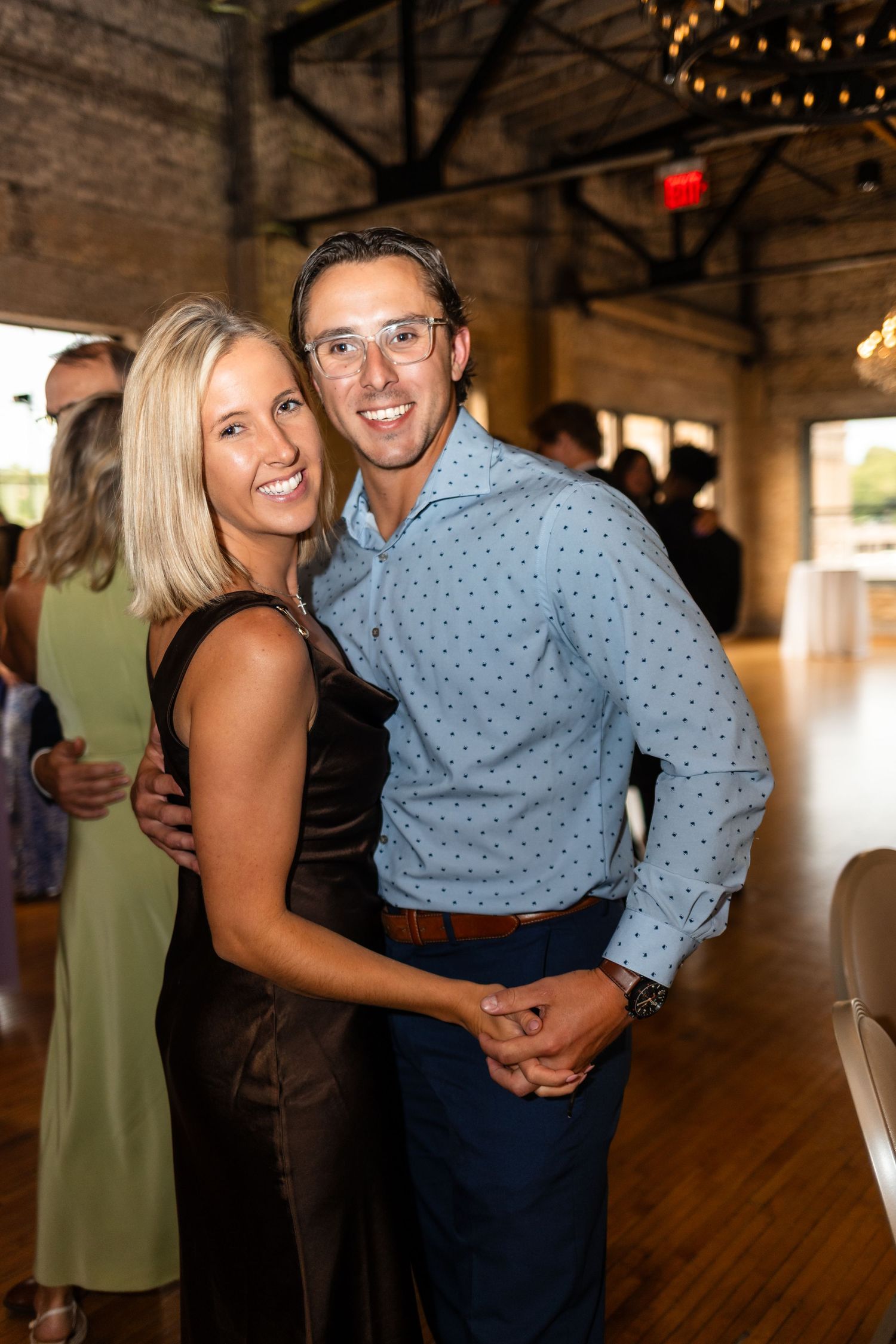 A smiling couple poses together at an indoor event, woman in dark dress and man in light blue shirt.