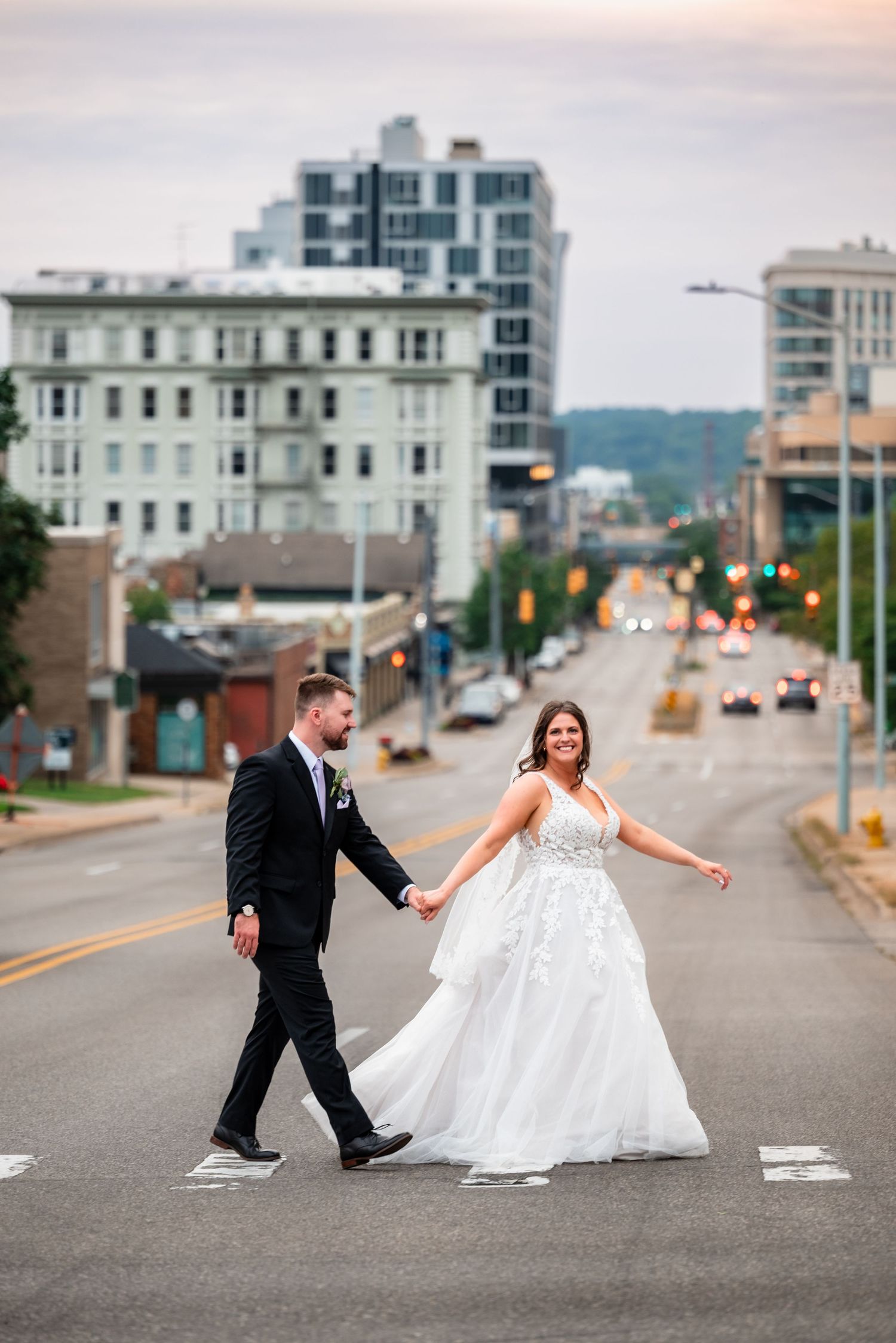 Bride and groom cross a city street at dusk, holding hands, bride in white gown, groom in black suit.