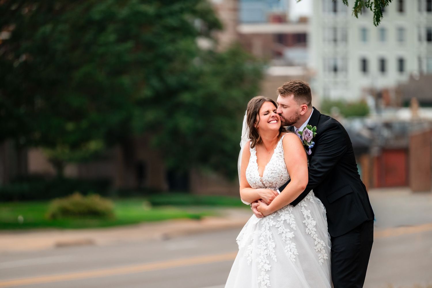 Bride and groom share a romantic embrace on a city street, with the groom kissing her cheek in wedding attire.