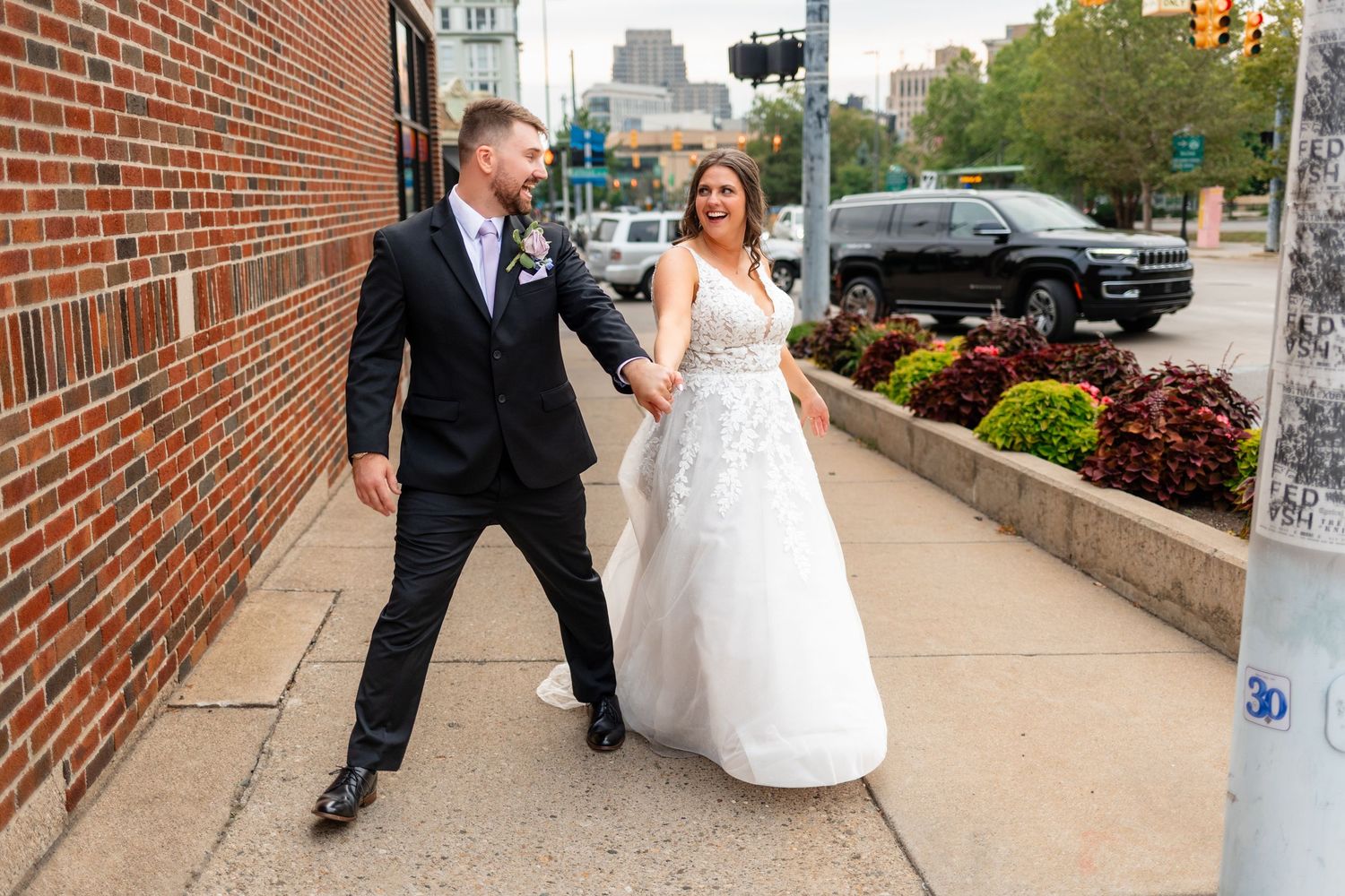 Bride and groom walk hand in hand along a city sidewalk, smiling joyfully in elegant wedding attire.