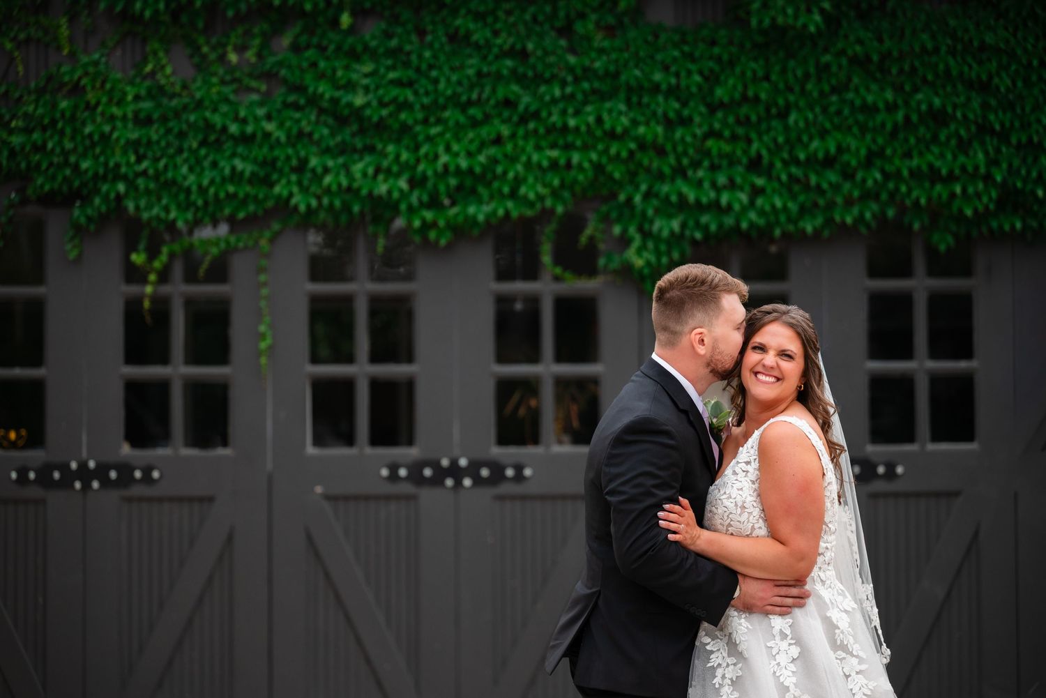 Bride and groom embrace romantically in front of gray barn doors covered with lush green ivy at outdoor wedding venue.