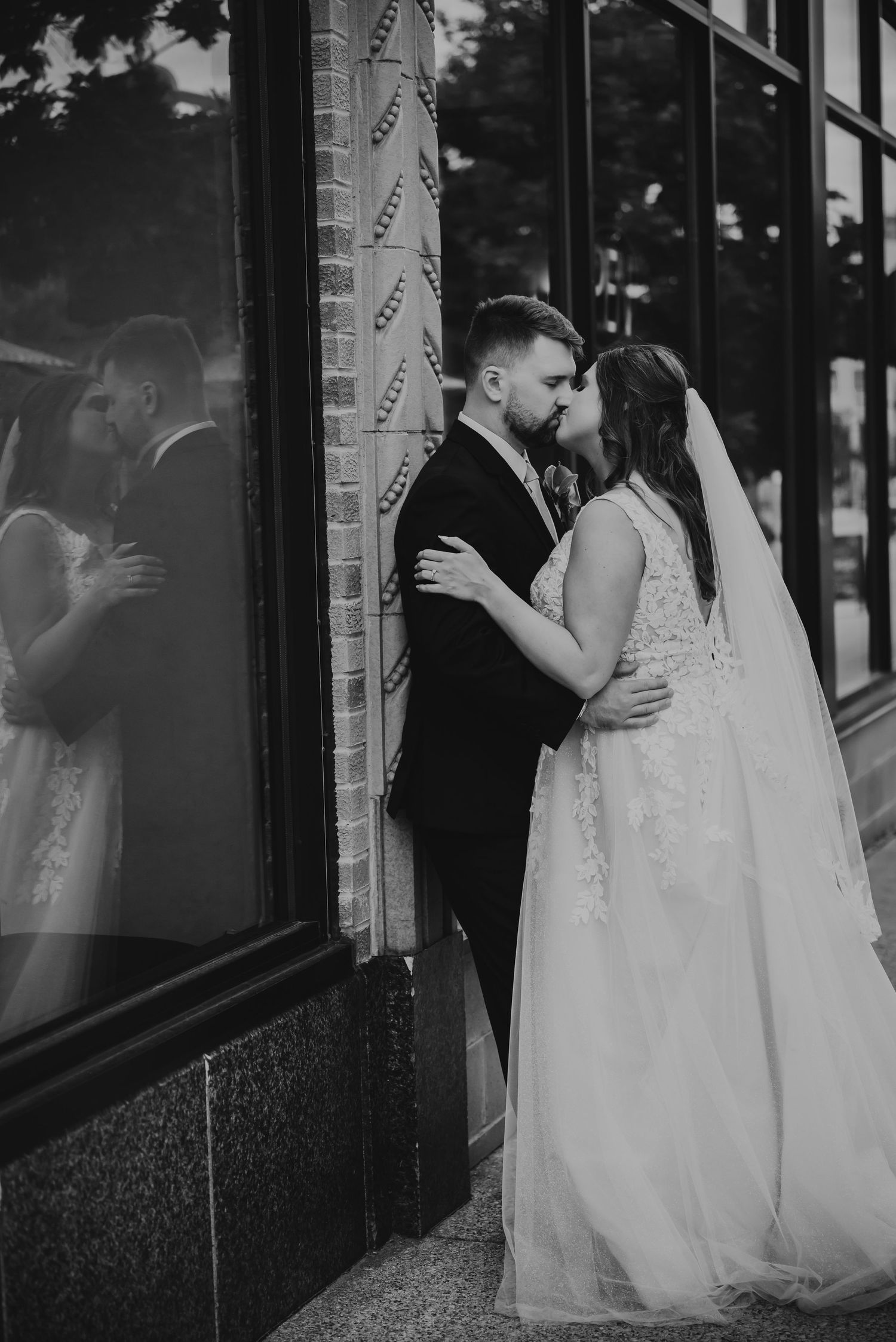 Black and white photo of a bride and groom kissing against a brick building, their reflection visible in the window.