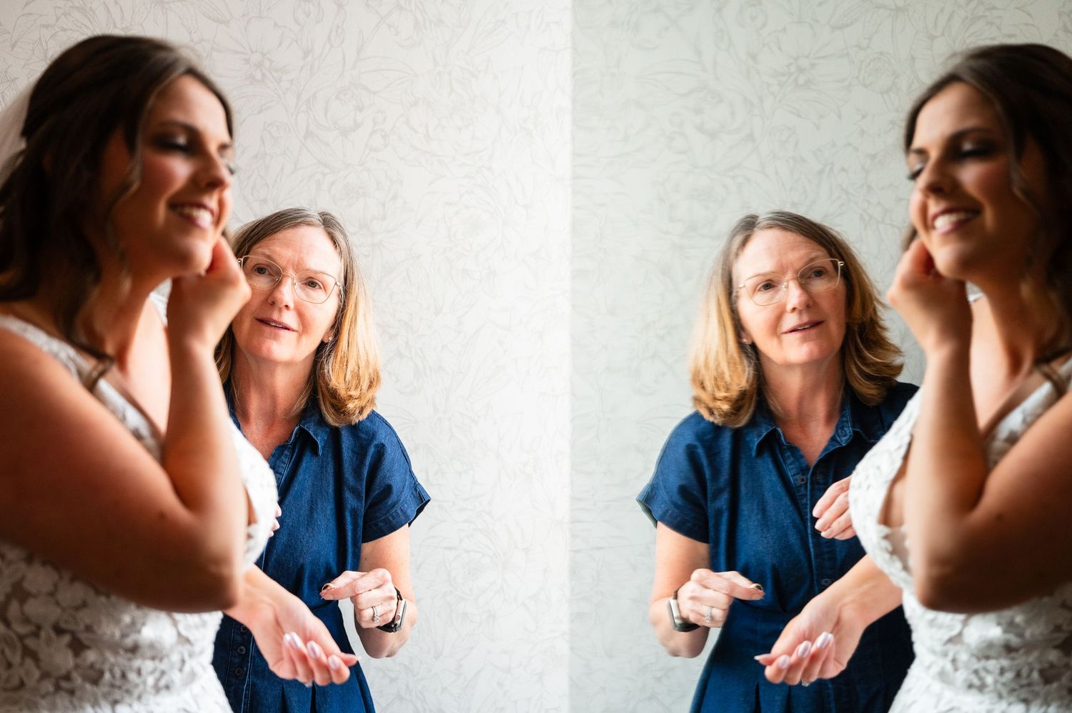 Bride putting on earrings with help from older woman in blue dress, reflected in mirror.