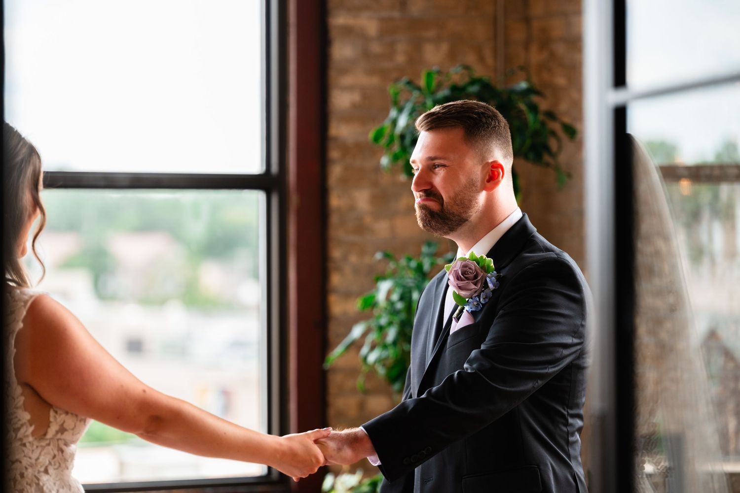 Groom in black suit with floral boutonniere holds bride's hands during emotional first look at wedding venue.