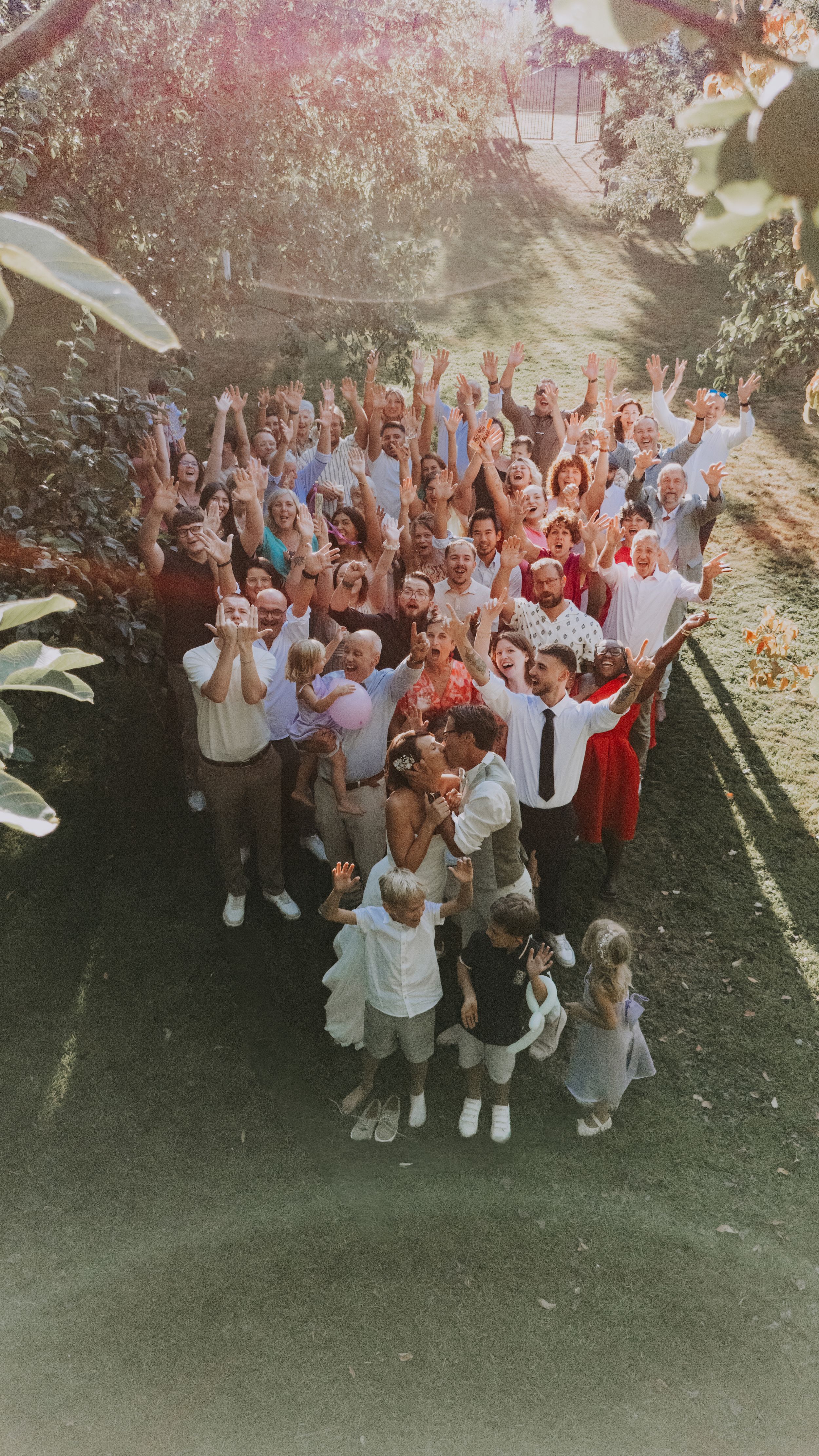 Large wedding group photo outdoors with guests cheering and raising hands, surrounded by lush greenery in warm sunlight.