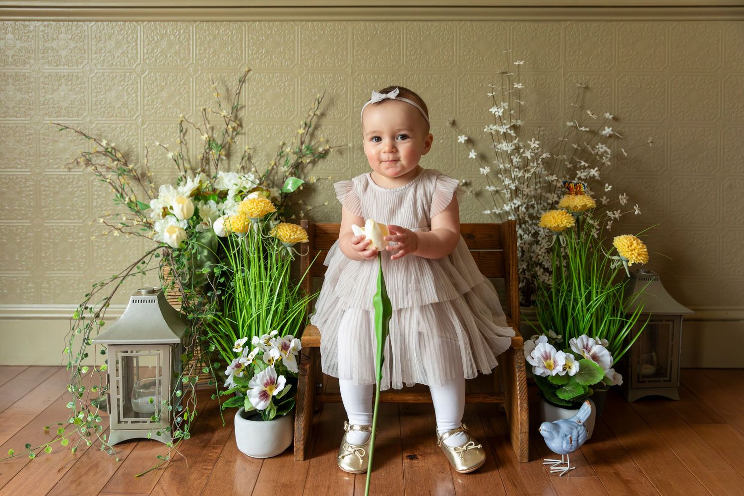 Baby girl in a white dress posing for birthday photos in Maine, surrounded by spring flowers and lanterns in a bright indoor setting.
