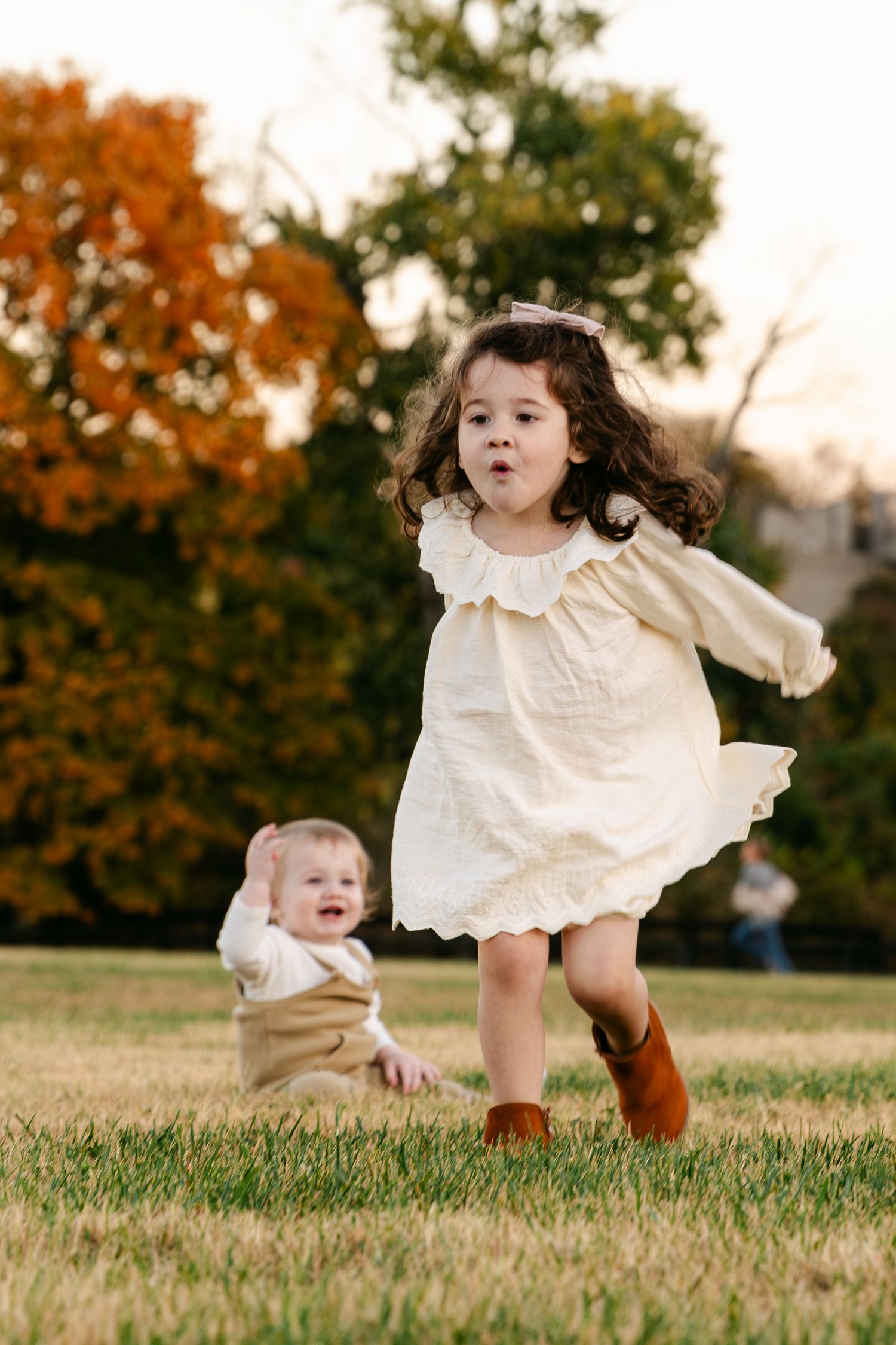 Two young children playing outdoors on grass in autumn, older girl in cream dress and boots running joyfully.