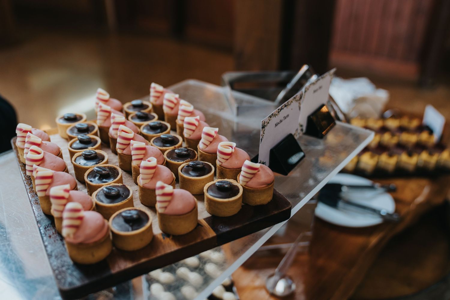 Elegant dessert display featuring chocolate tarts topped with pink cream swirls arranged on a wooden board at a catered event.