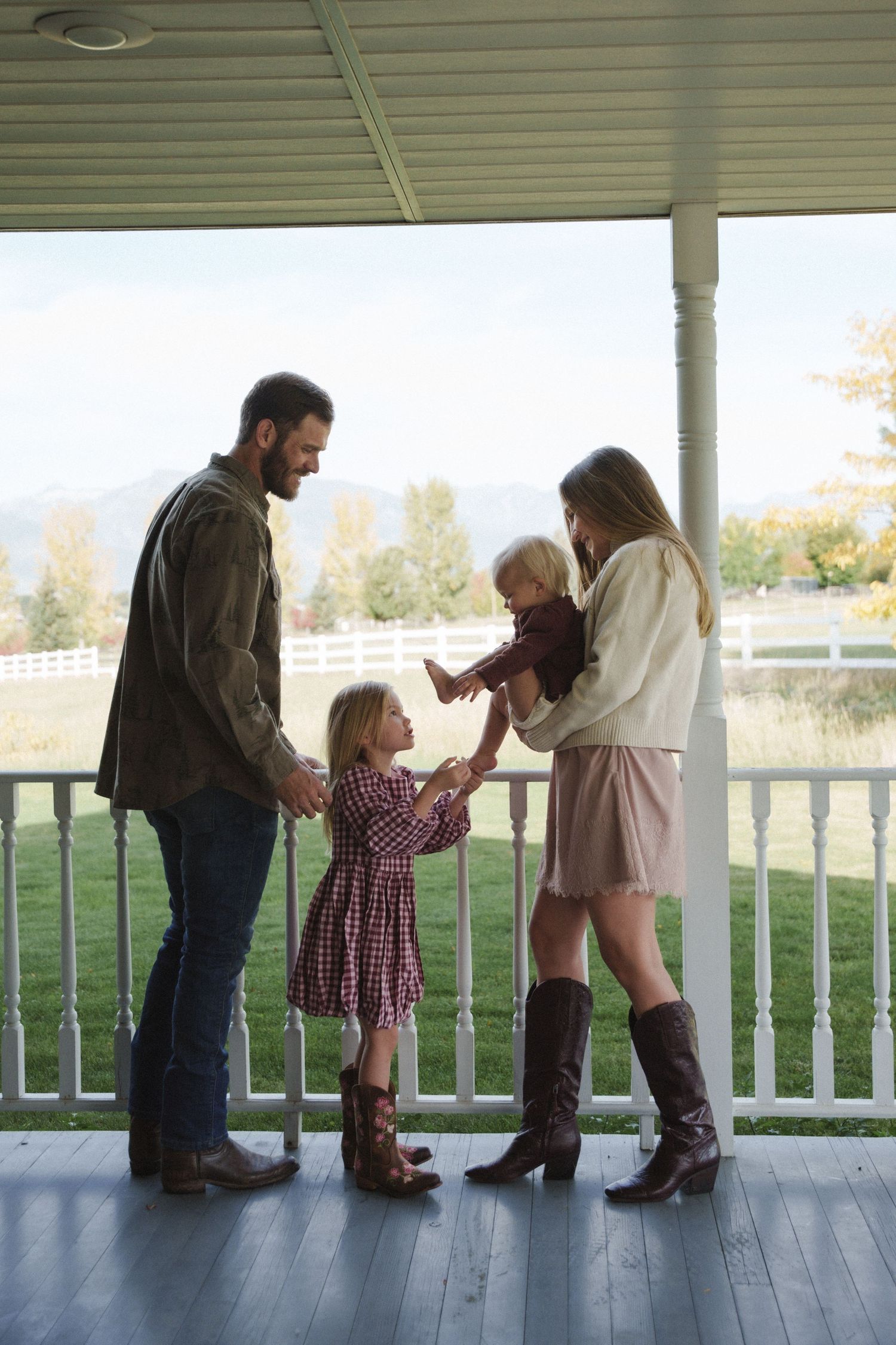 Family of four sharing a warm moment on a farmhouse porch, with white fence and mountains in background.