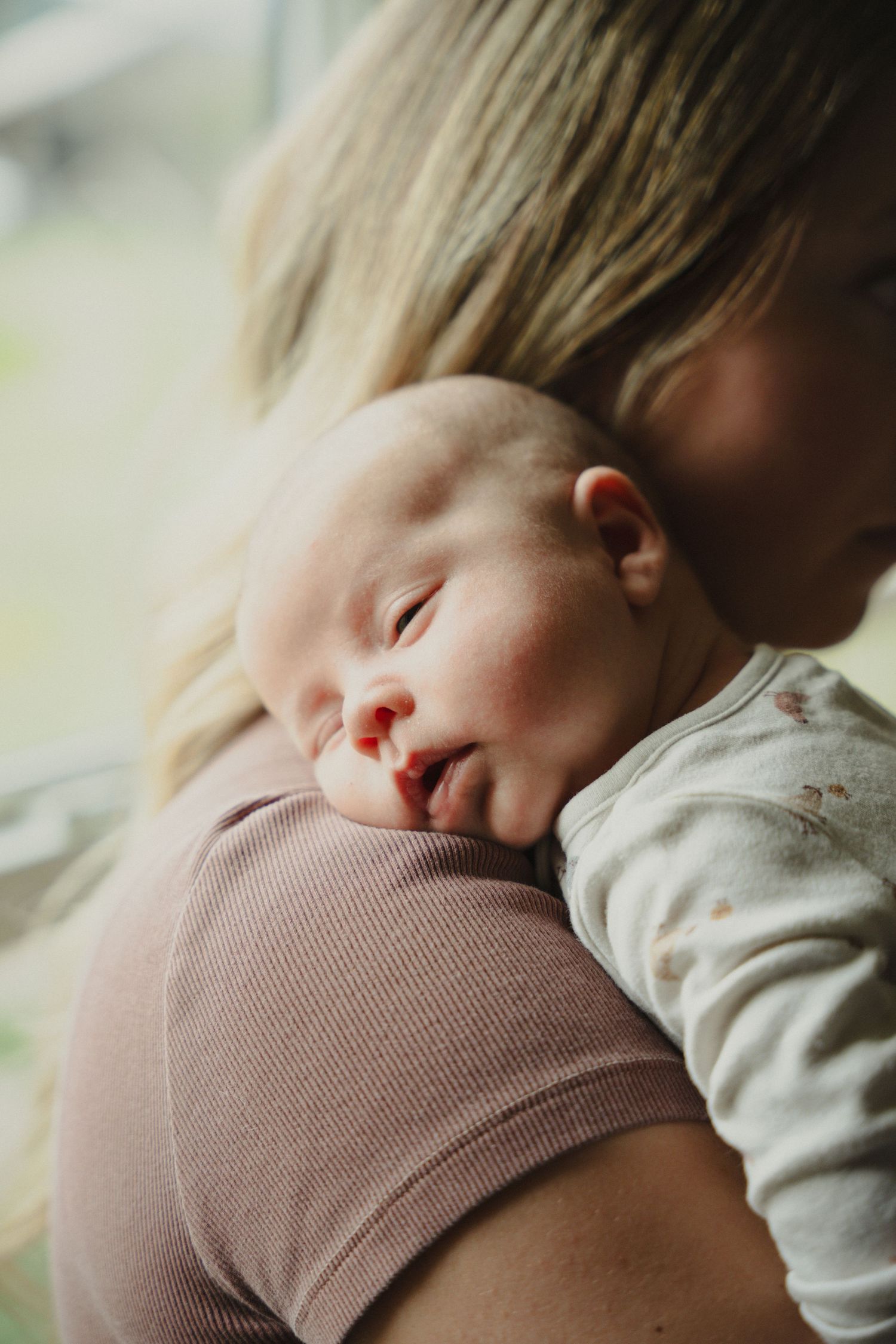 A newborn baby sleeping peacefully on a mother's shoulder, cradled in her arms near a window.