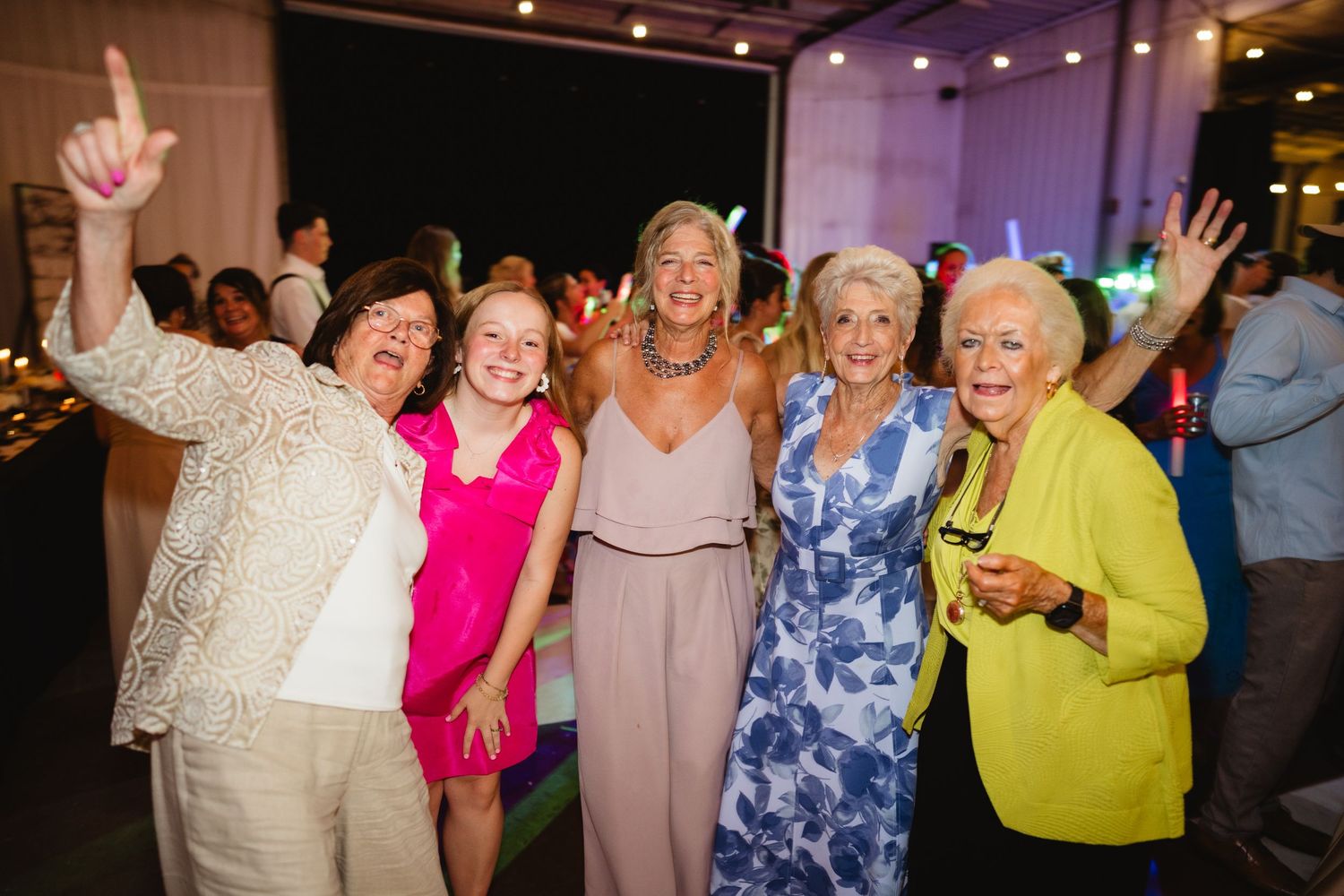 Five women smiling and posing together at a festive indoor party event, one raising her hand in a shaka sign.