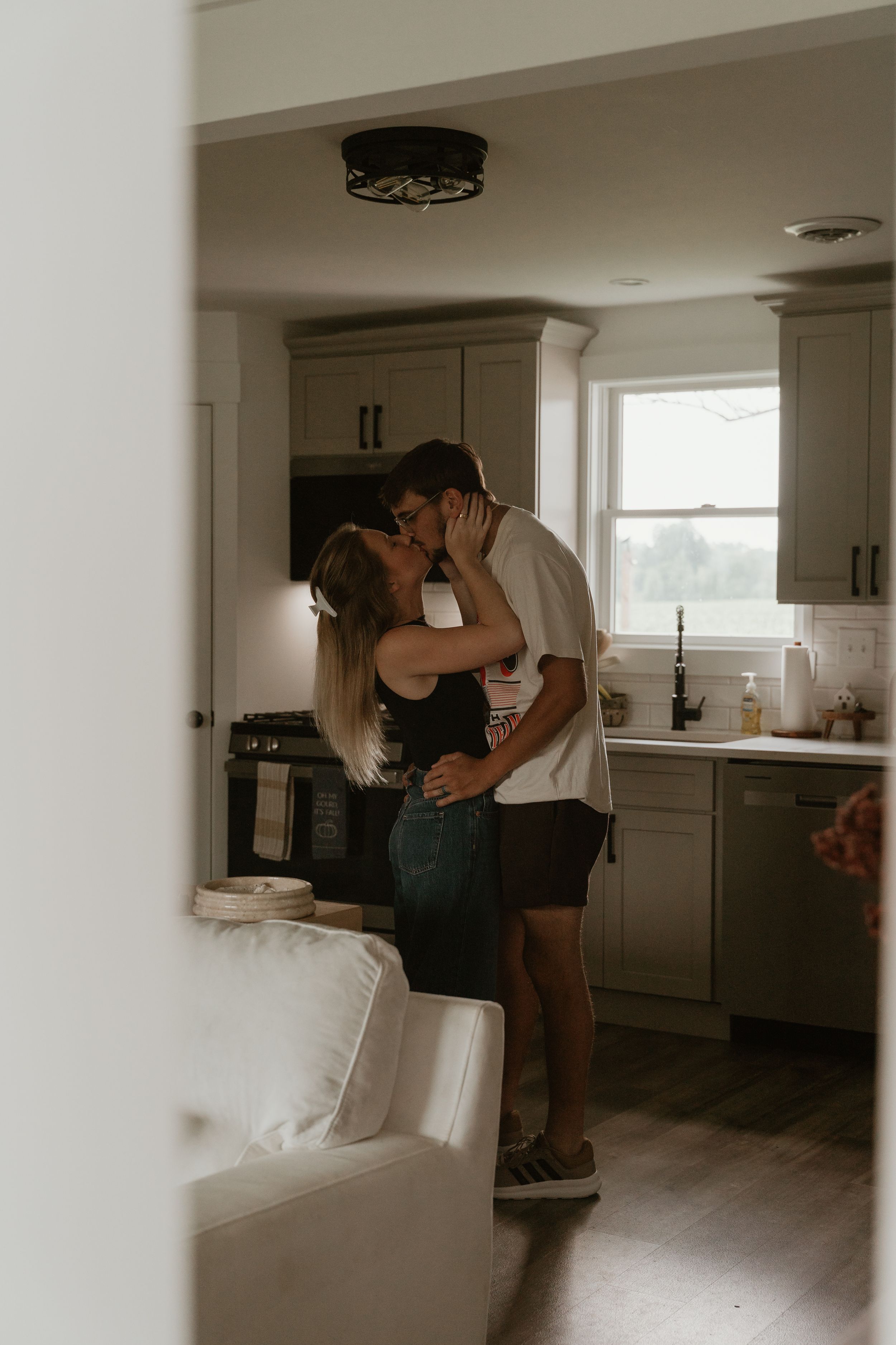 A couple sharing a romantic kiss in a cozy kitchen with sage green cabinets and hardwood floors.