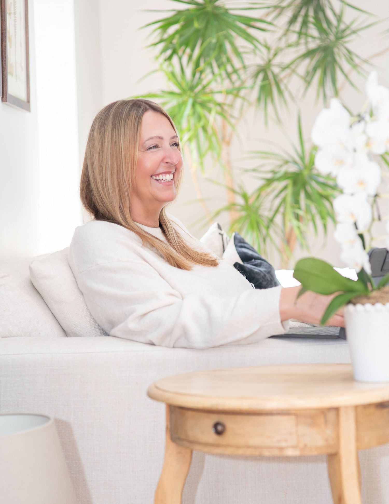 Smiling blonde woman in white blazer using laptop on couch beside orchid and tropical plants in bright living room.