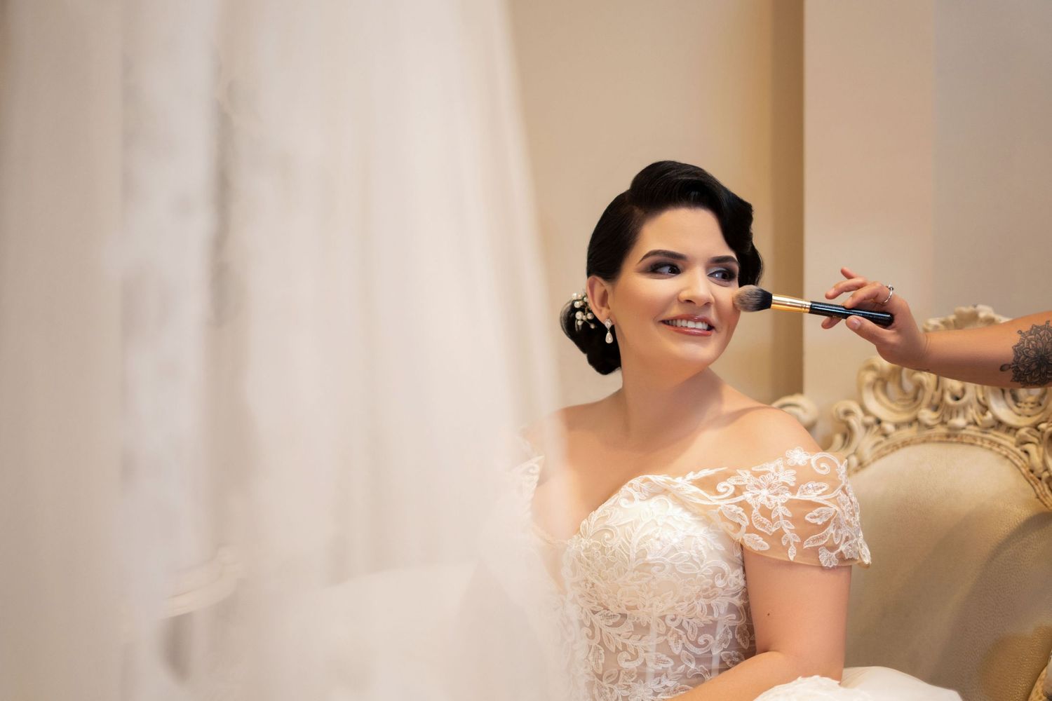 Bride in lace wedding dress getting makeup applied by artist while smiling in elegant chair.