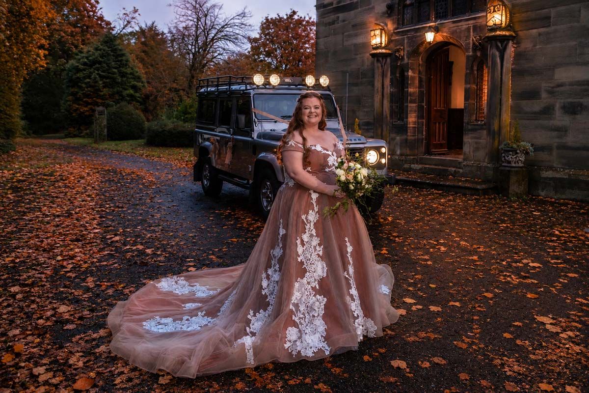 Bride in champagne floral gown poses with Land Rover amid autumn leaves at dusk outside stone manor.