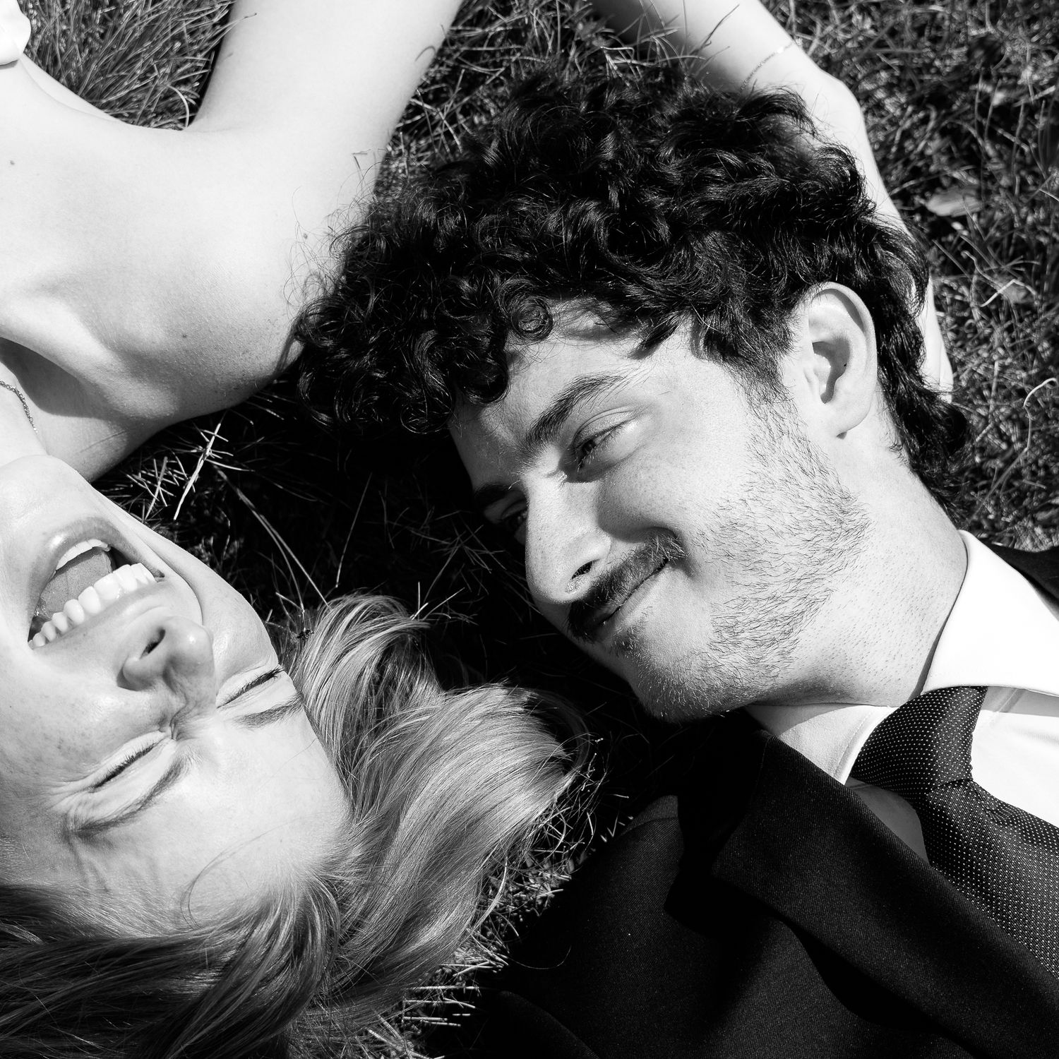 Black and white photo of a smiling couple lying on grass, gazing at each other in a romantic outdoor setting.