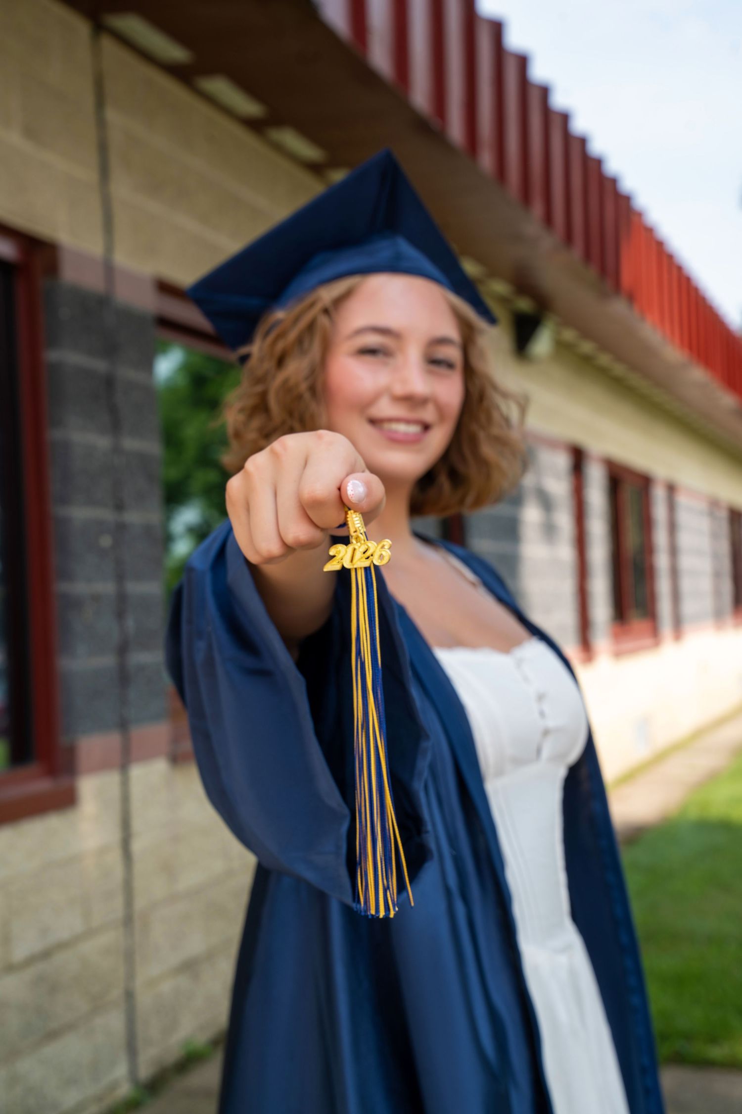 Smiling graduate in navy cap and gown holds 2024 tassel toward camera outside school building.