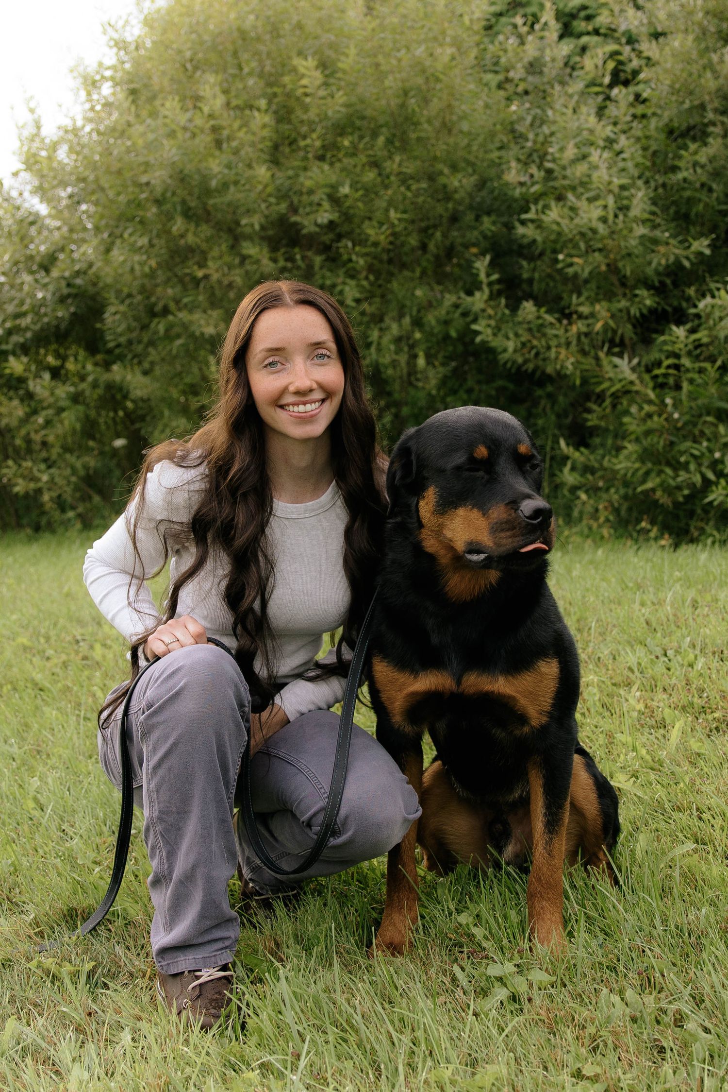 Smiling young woman with long brown hair kneeling beside a Rottweiler dog on green grass outdoors.