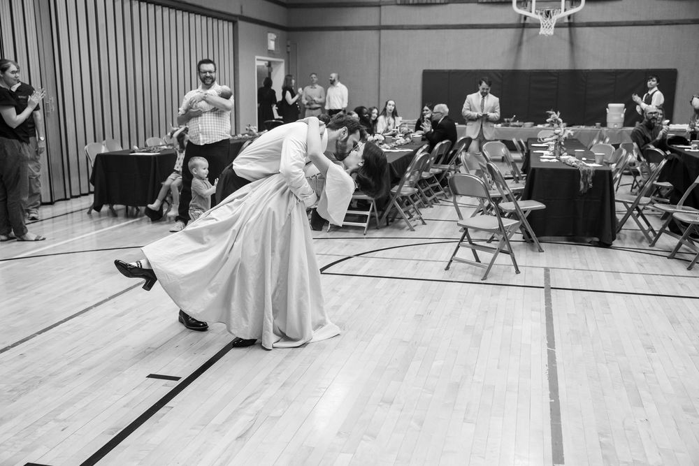 Couple shares romantic dip dance on gymnasium floor during wedding reception in black and white photo.