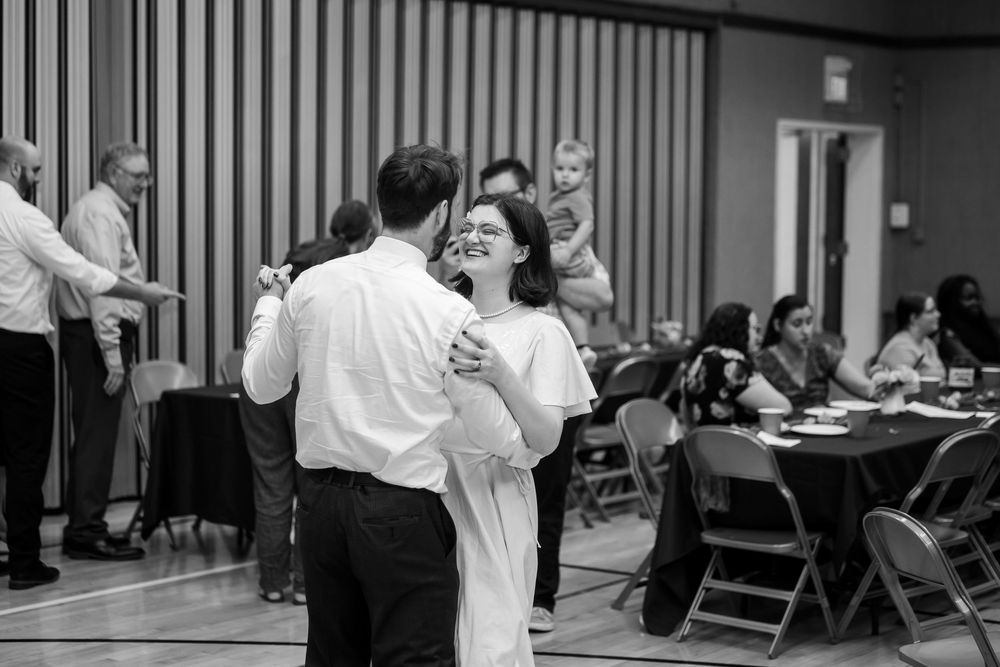 Couple sharing a romantic dance at a formal event in a decorated hall with guests seated at tables in background.
