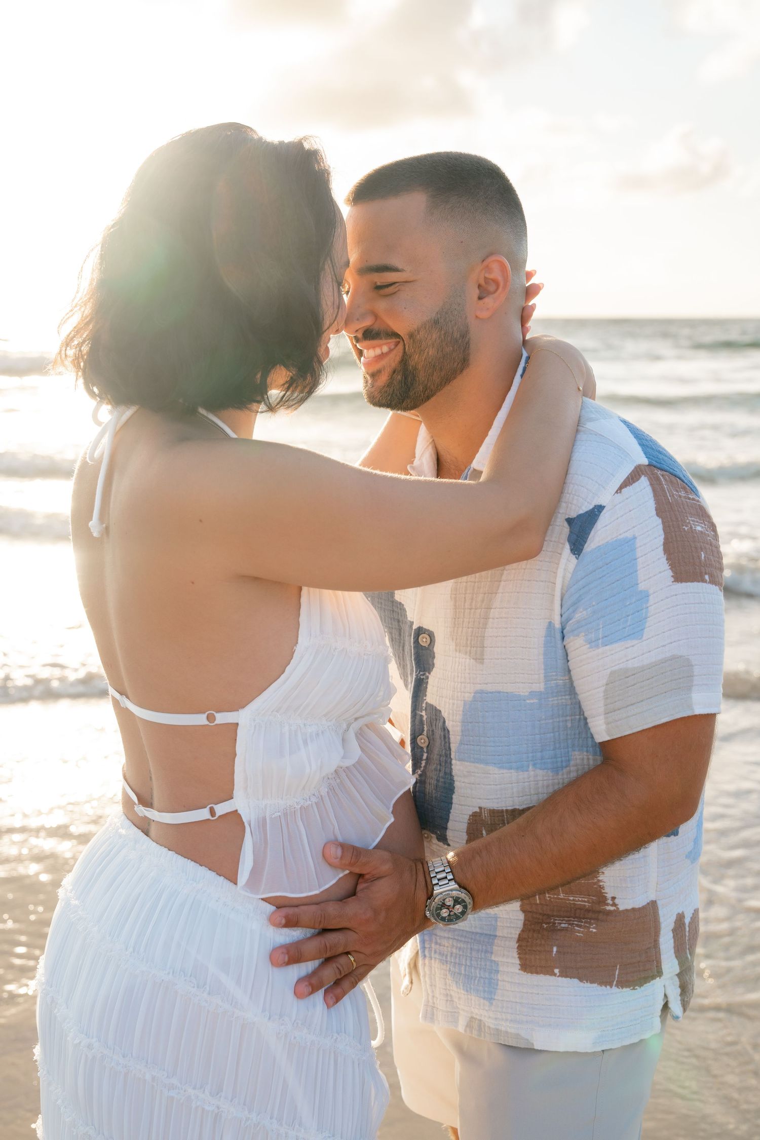Smiling couple embracing on a sunny beach, woman in white dress, man in patterned shirt, ocean waves behind them.