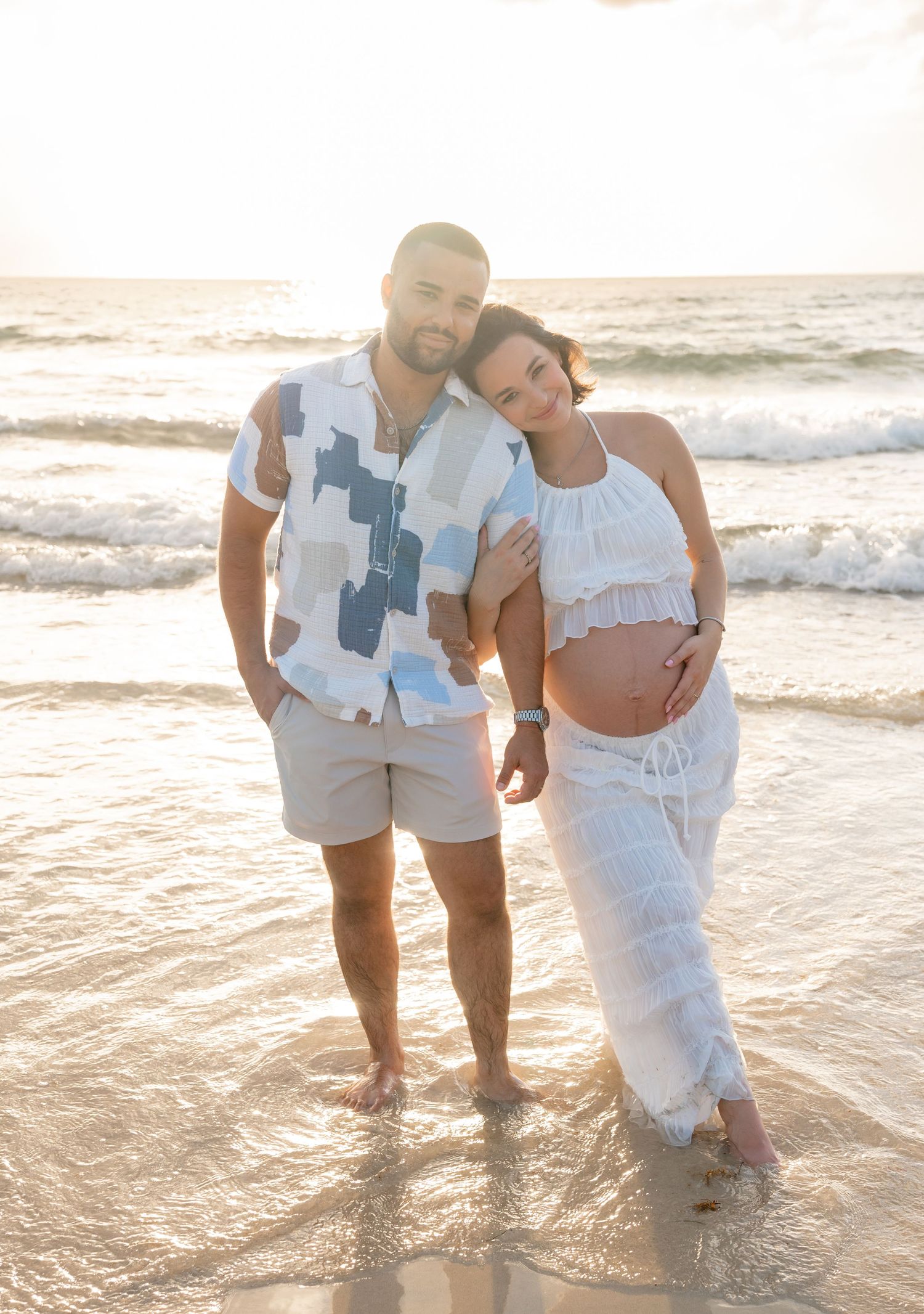 Pregnant couple posing on a beach at sunset, man in patterned shirt and shorts, woman in white maternity outfit.