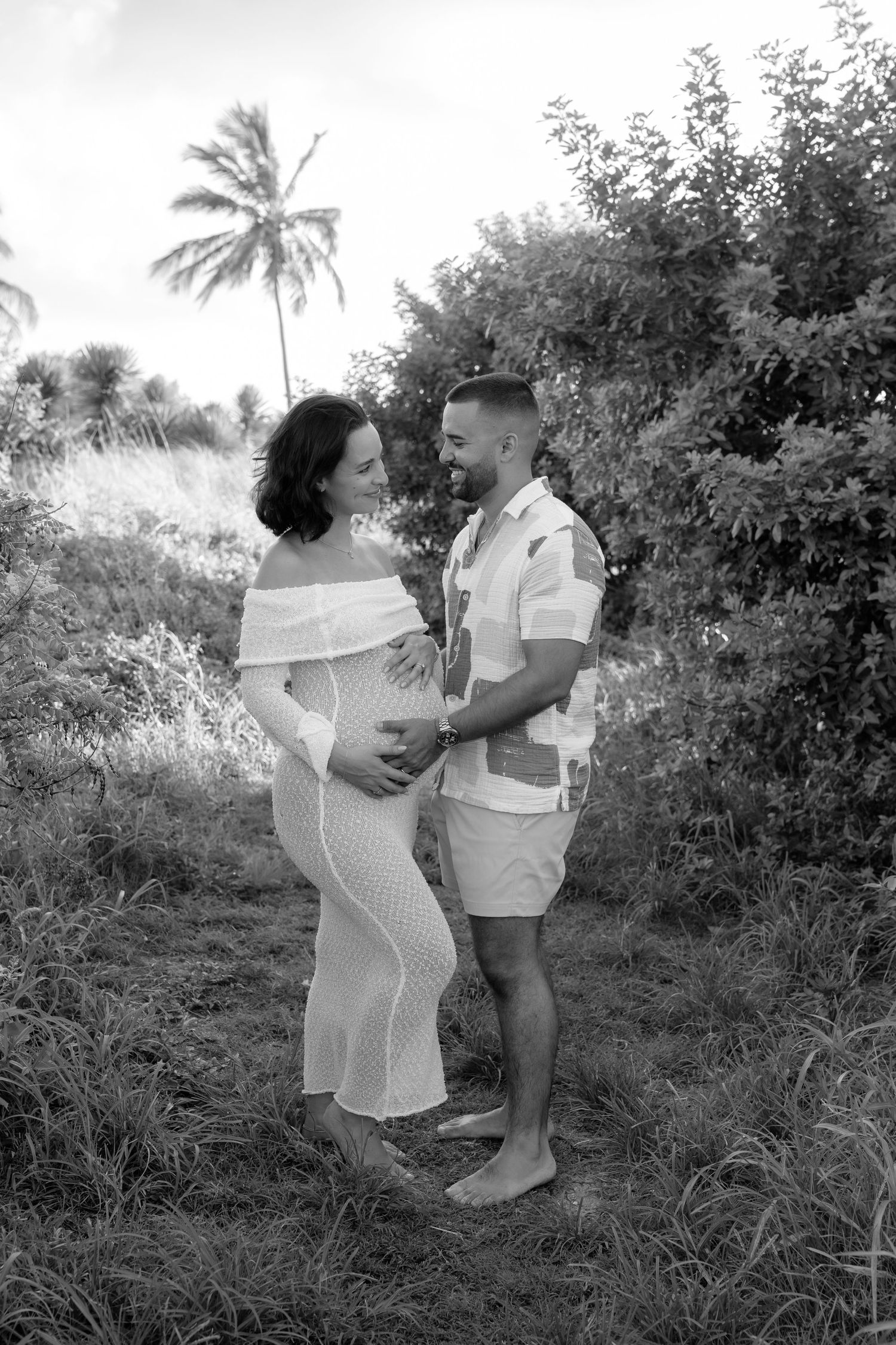 Black and white maternity photo of a couple smiling at each other outdoors with tropical trees in the background.