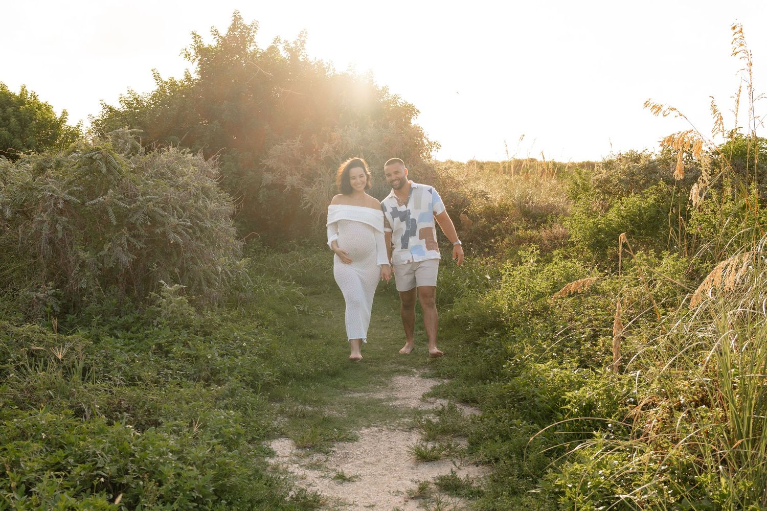 Couple walking hand-in-hand on a sunlit nature path surrounded by lush green bushes during golden hour.