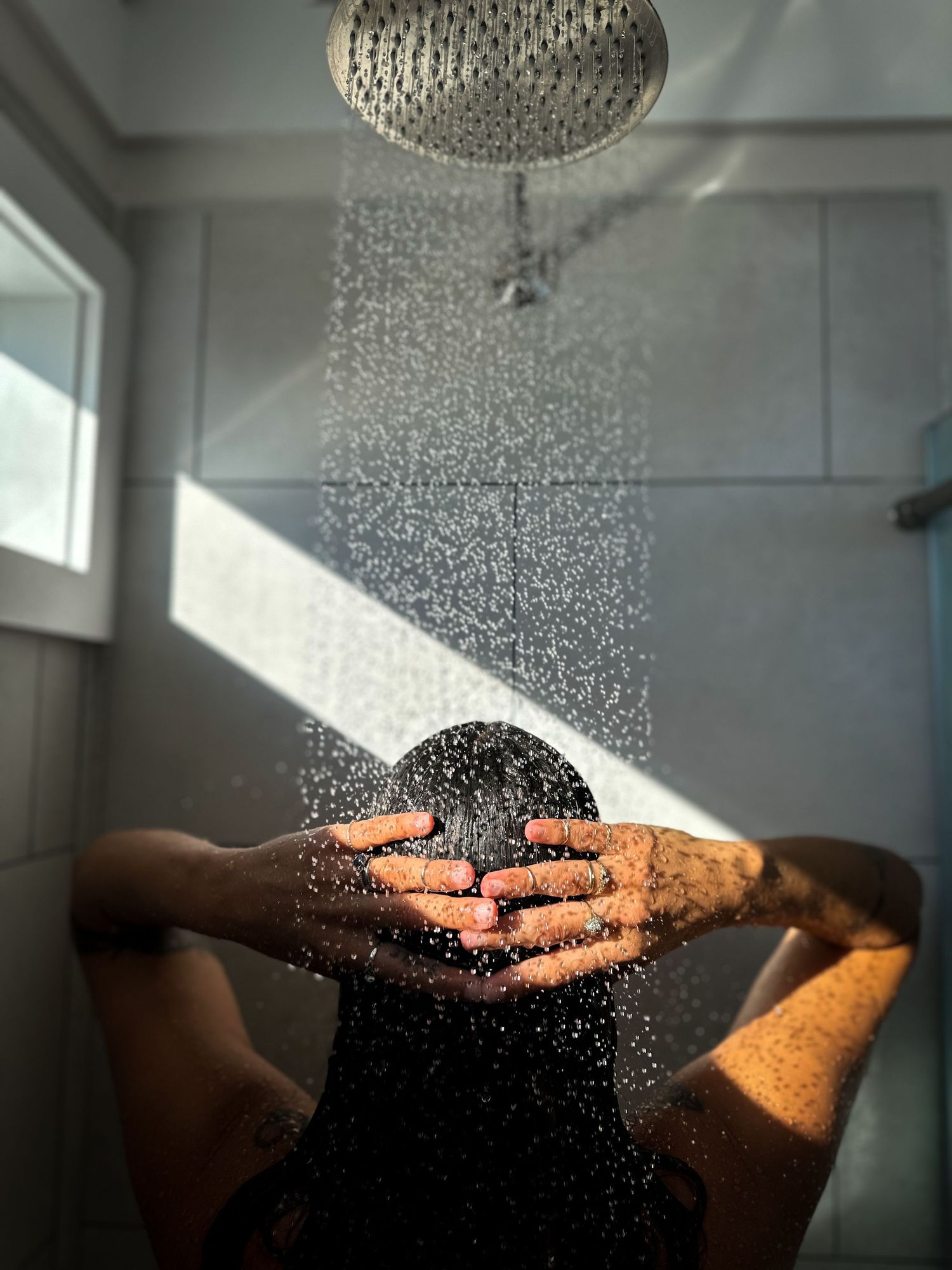 Person showering under large rain showerhead with sunlight streaming through bathroom window.