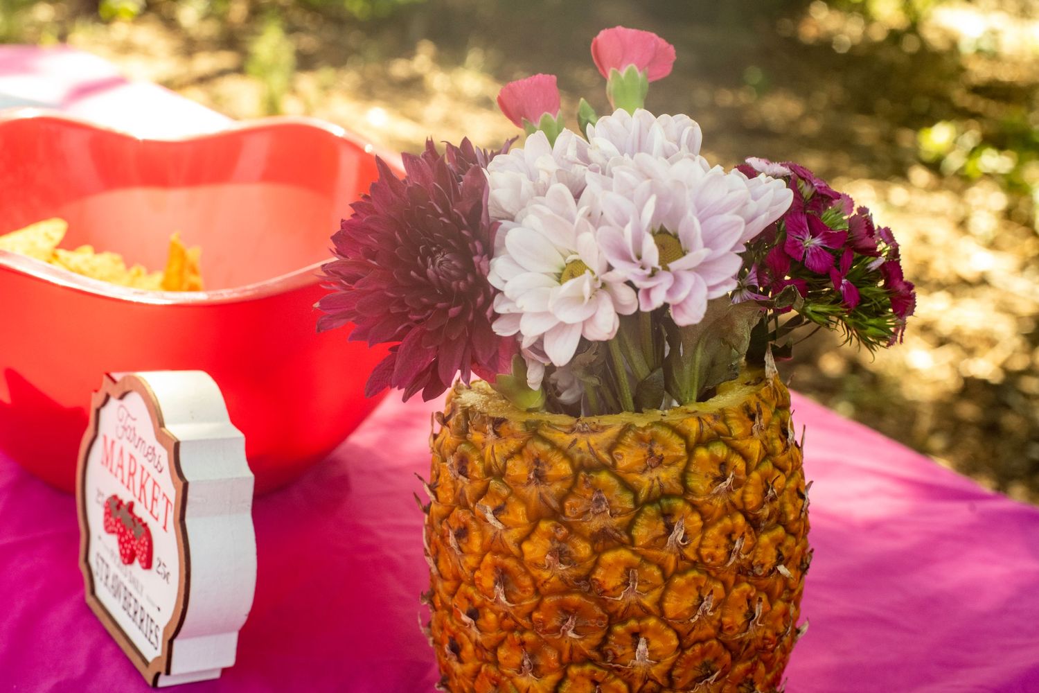 Floral arrangement in a hollowed pineapple vase on a pink tablecloth at an outdoor party with a red bowl nearby.