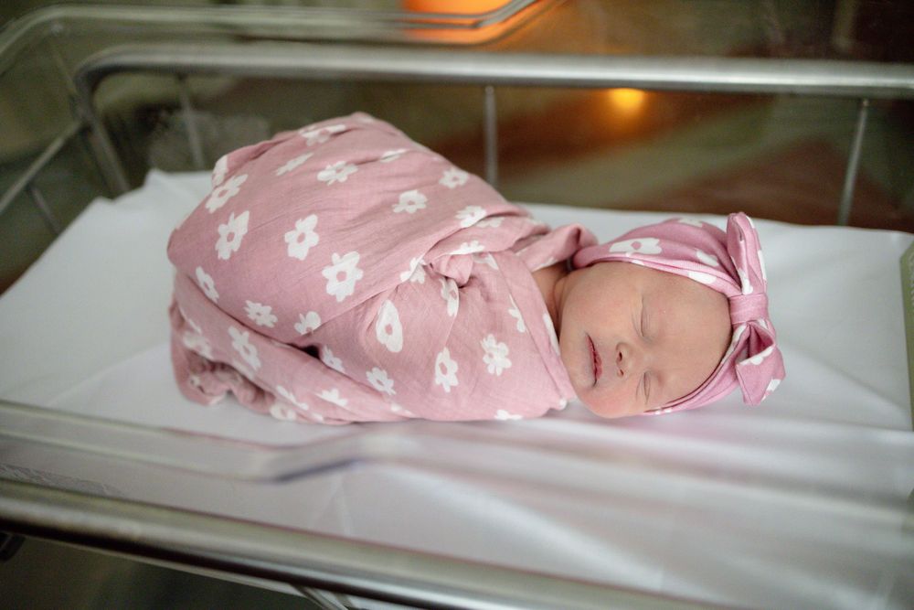 Newborn baby girl swaddled in pink floral blanket with matching bow headband sleeping peacefully in hospital bassinet at Parkwest.