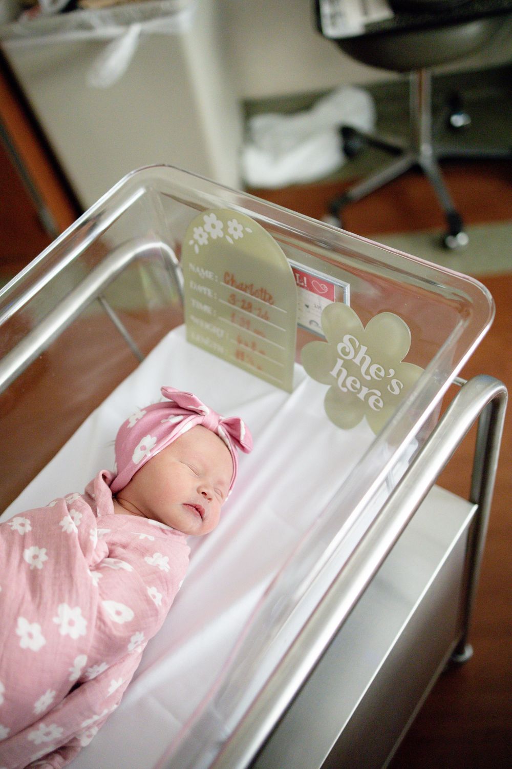 Newborn baby girl swaddled in pink floral blanket sleeping peacefully in hospital bassinet with She's Here sign at Parkwest Medical Center.