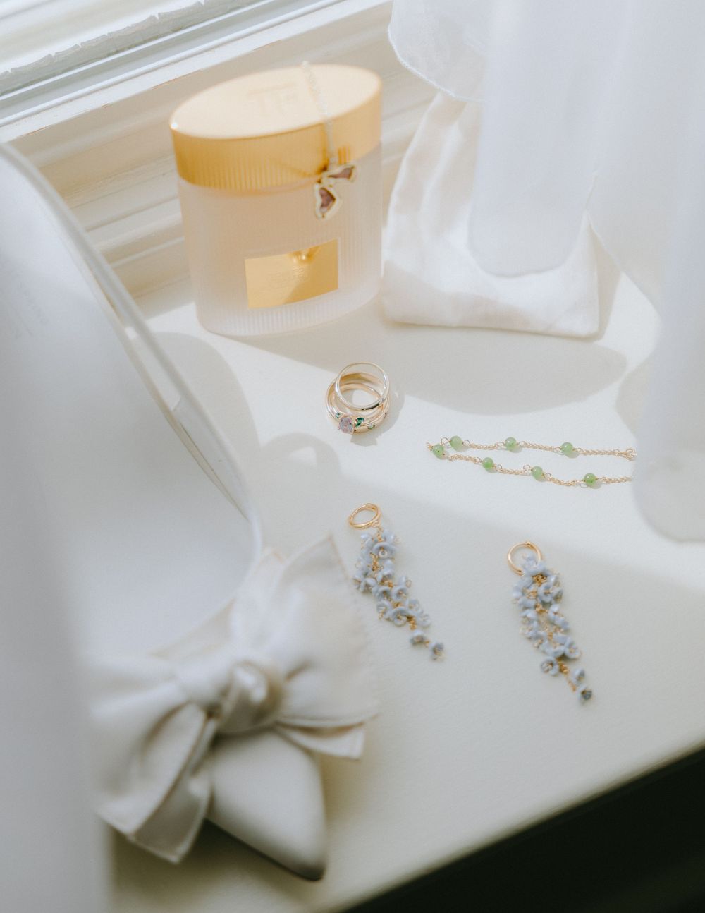 Elegant bridal flat lay with gold rings, floral earrings, green bead necklace, and a yellow gift box on white satin.