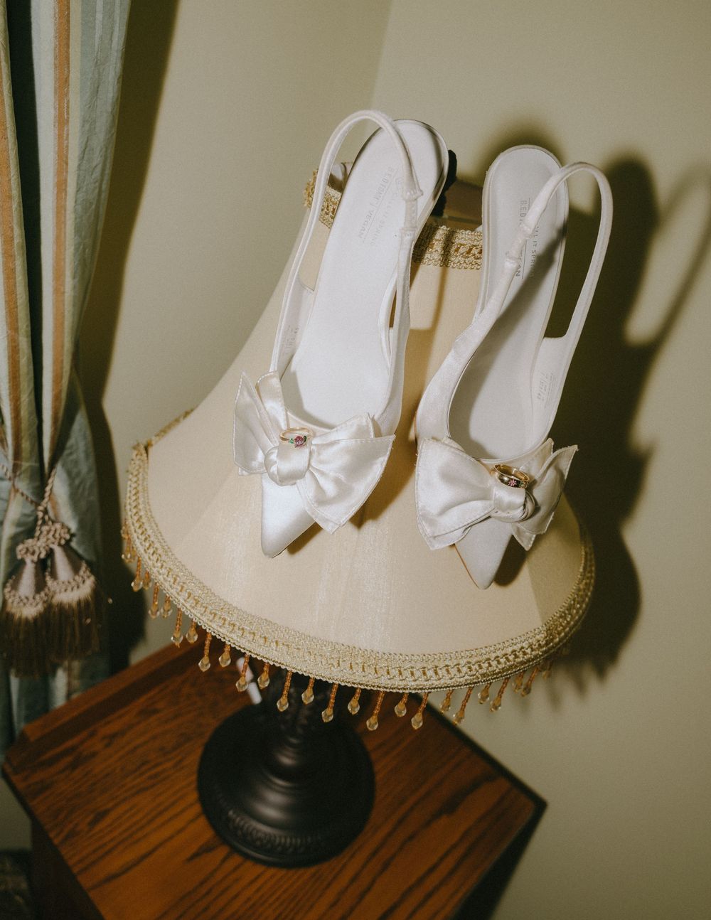 White satin bridal heels with bow detail displayed on a vintage lamp shade in an elegant room setting.