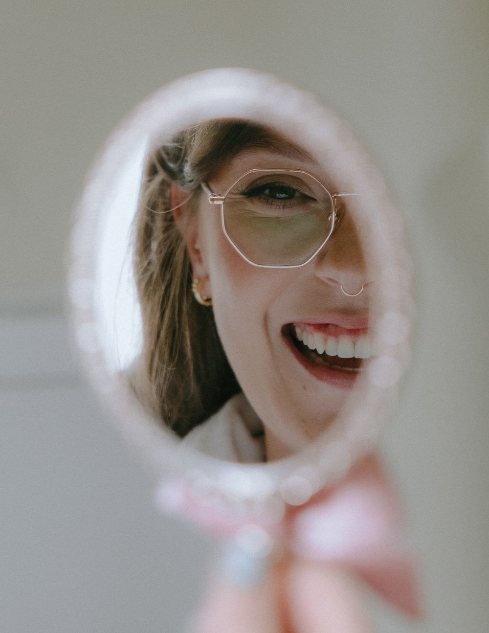 A smiling woman with geometric glasses and a nose ring reflected in a small oval handheld mirror.