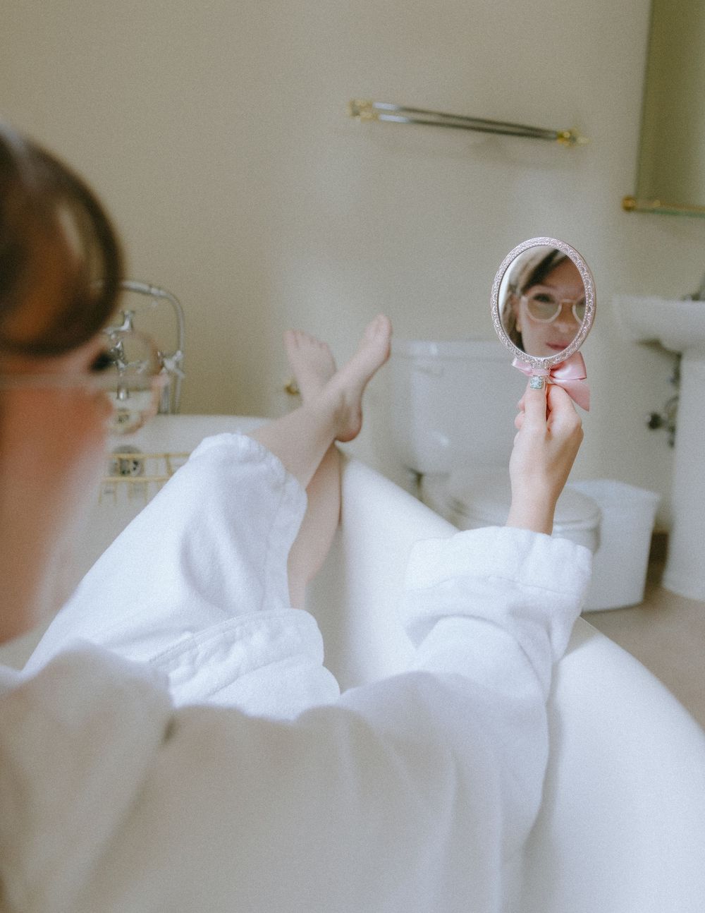 Woman in white robe relaxing in bathtub holding small round mirror reflecting her face.