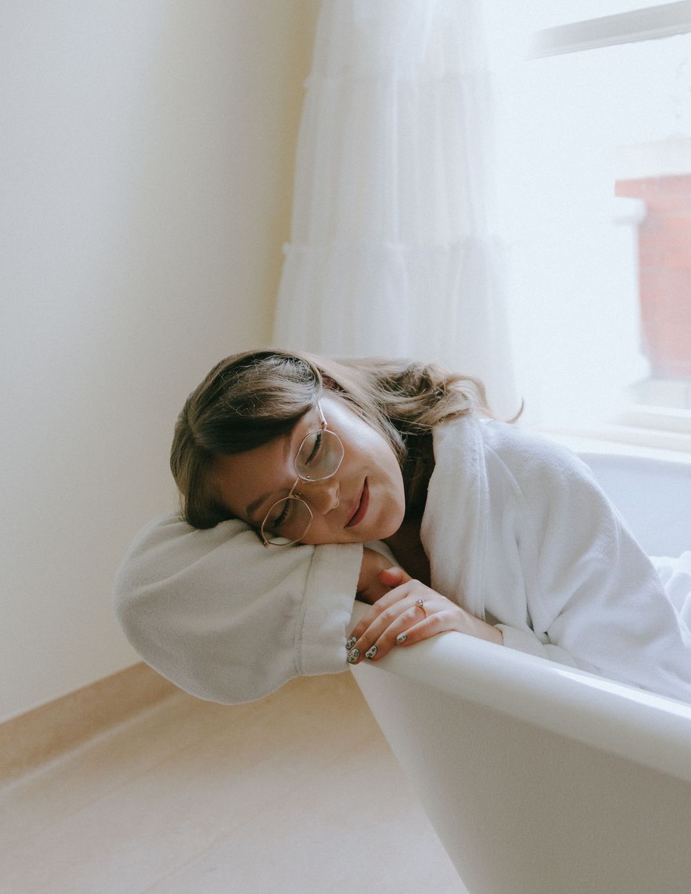 Woman in white robe resting on bathtub edge near window with sheer curtains, wearing clear-framed glasses.