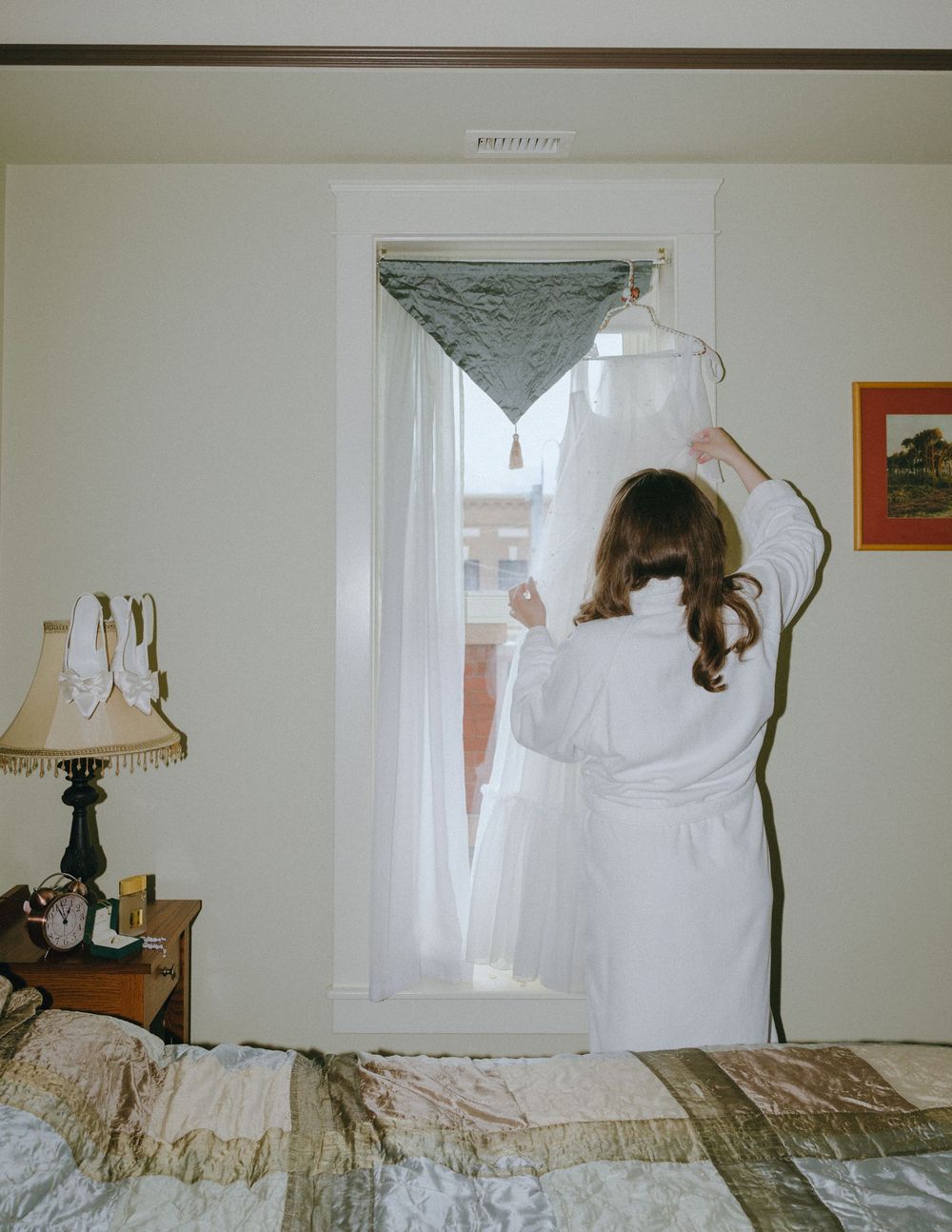 A bride in a white robe holds her wedding dress on a hanger near a window in a cozy hotel room.