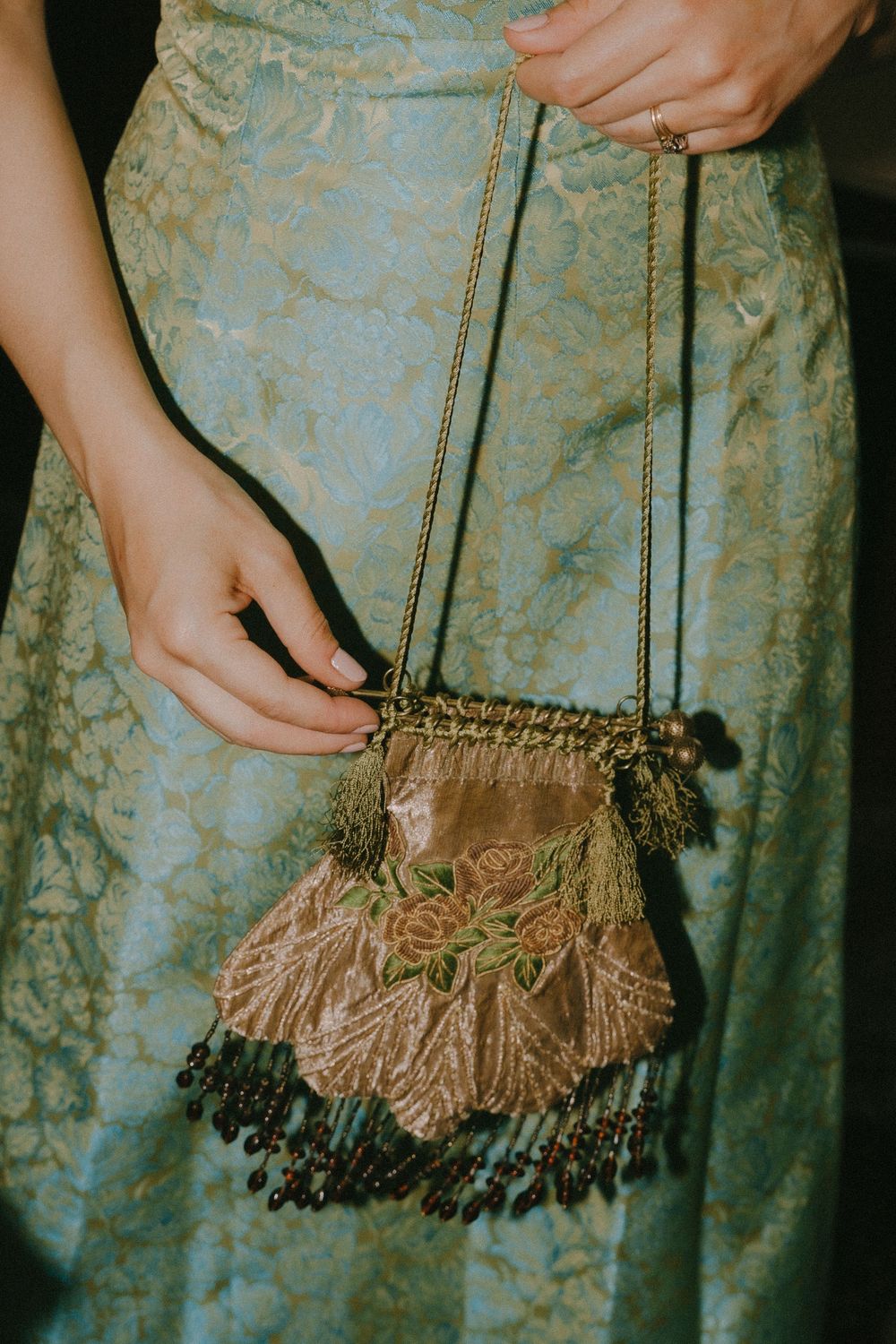 Woman in blue brocade dress holding vintage beaded purse with rose embroidery, tassels, and gold chain strap.