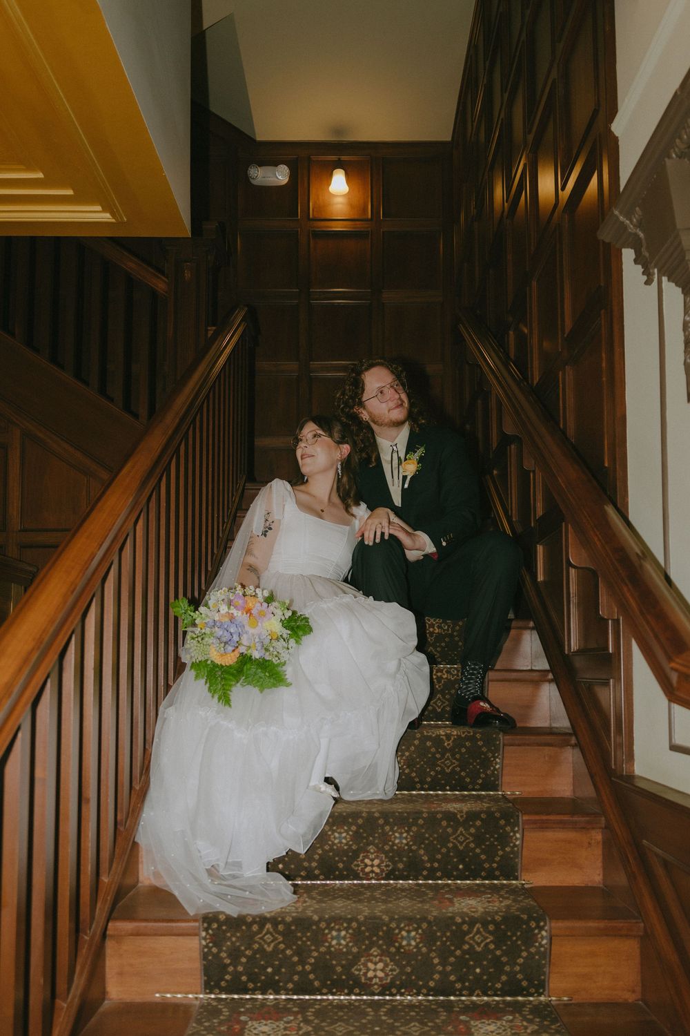 Bride and groom sitting together on elegant wooden staircase with colorful wedding bouquet.