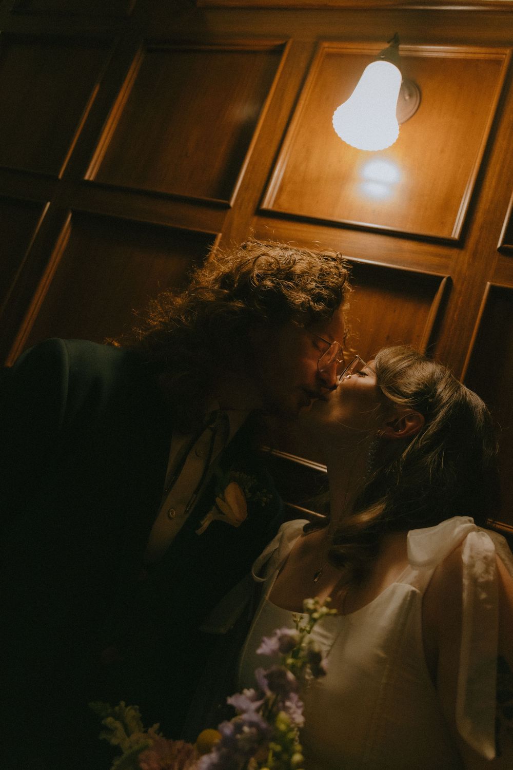 Couple sharing a romantic kiss under warm light, surrounded by flowers, in a wood-paneled room.