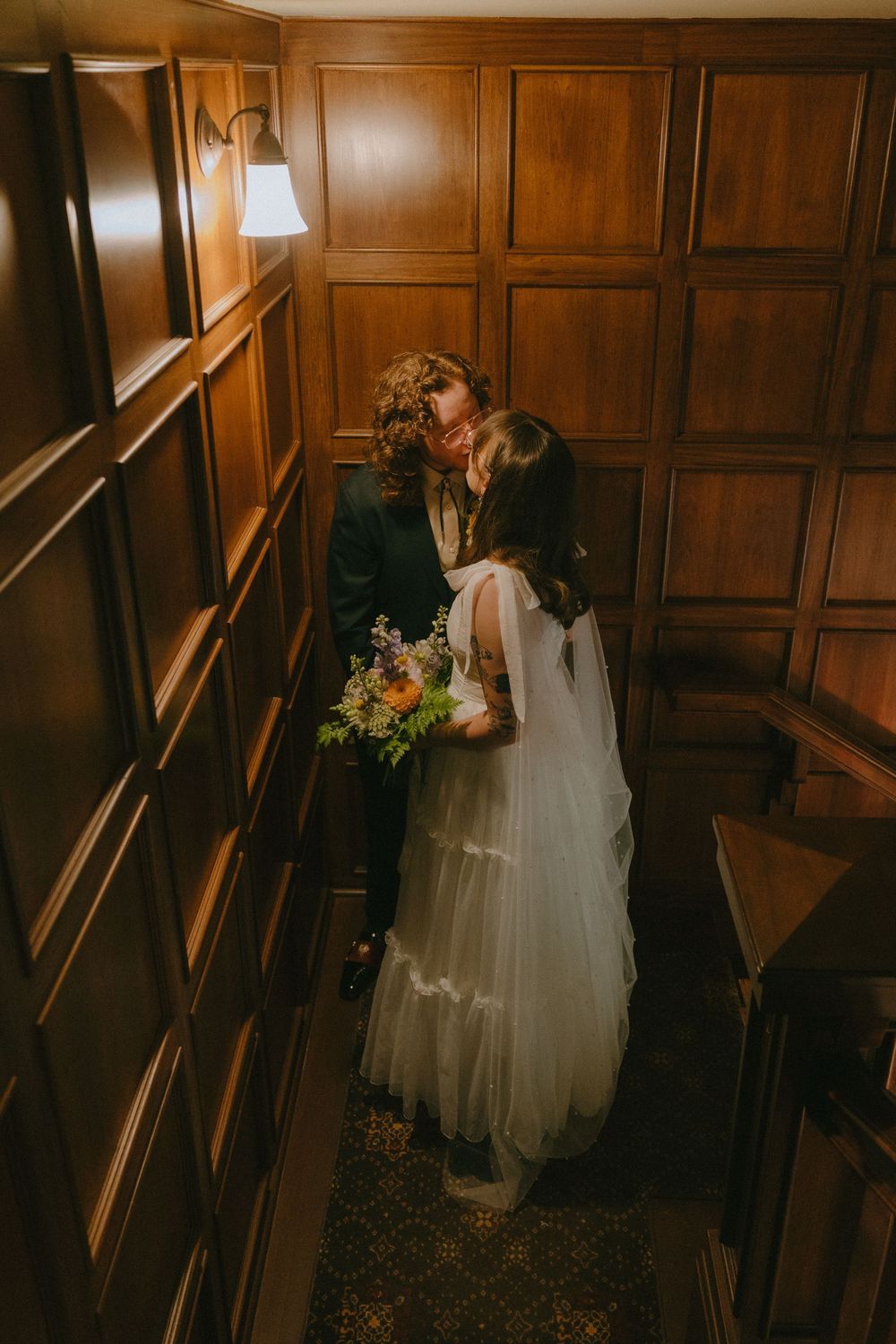 Bride and groom share a romantic kiss in a wood-paneled hallway, bride holding a colorful bouquet in a tiered white gown.